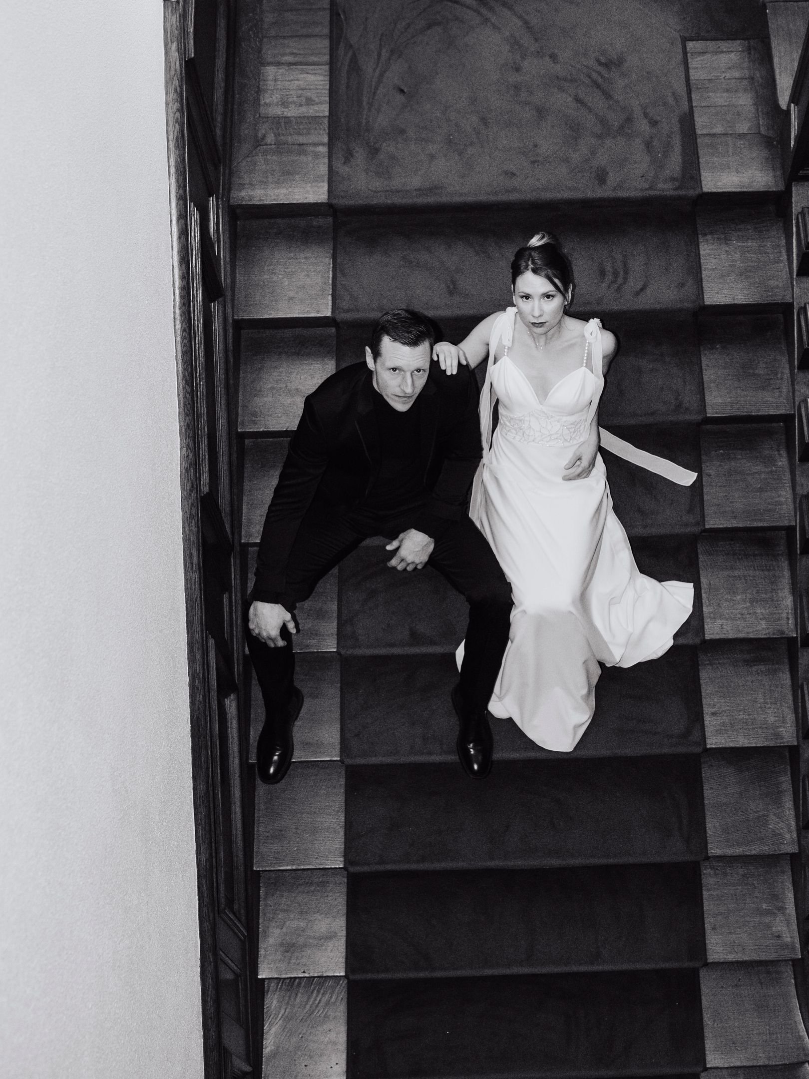 Une femme en robe de mariée et un homme en costume noir, vus du dessus, assis sur un escalier en bois