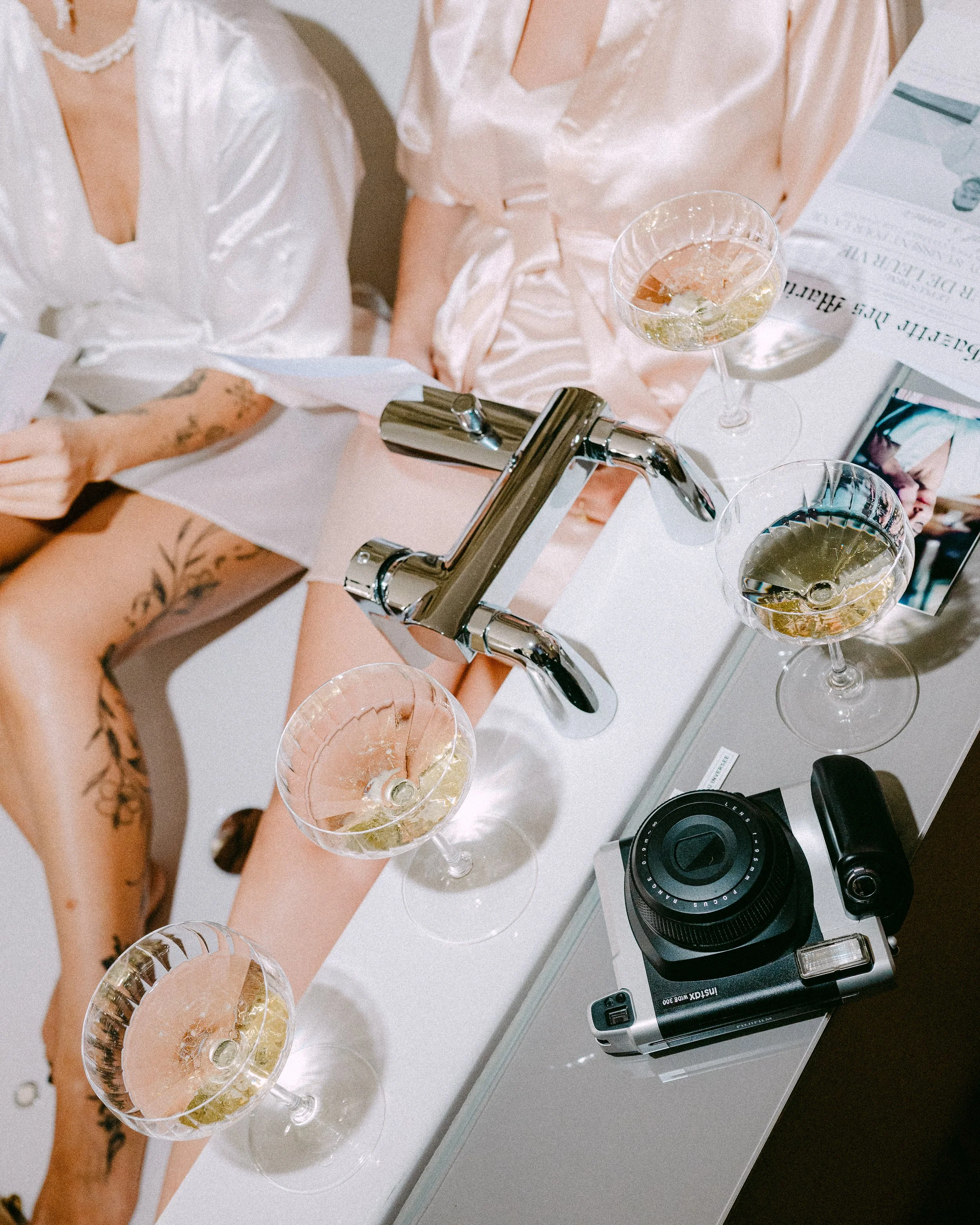 Gros plan sur un évier en chrome avec quatre verres à vin remplis de champagne. Deux personnes en robes de satin clair sont assises à côté, l'une avec tatouages visibles sur le bras et la cuisse. Sur la table à côté, il y a une vieille appareil photo