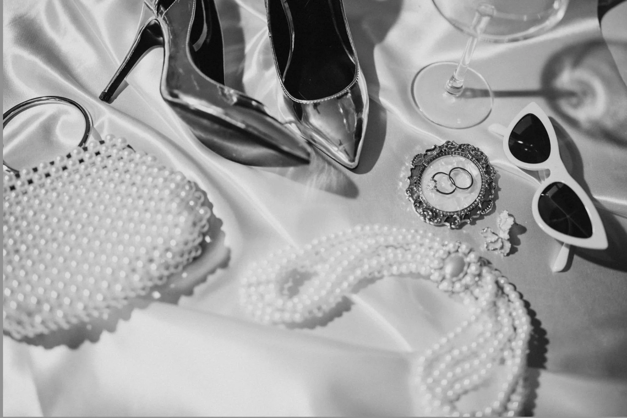 Barbie-themed accessories including pearl jewelry, high-heeled shoes, sunglasses, a mirror with rings, and a wine glass, arranged on satin fabric.