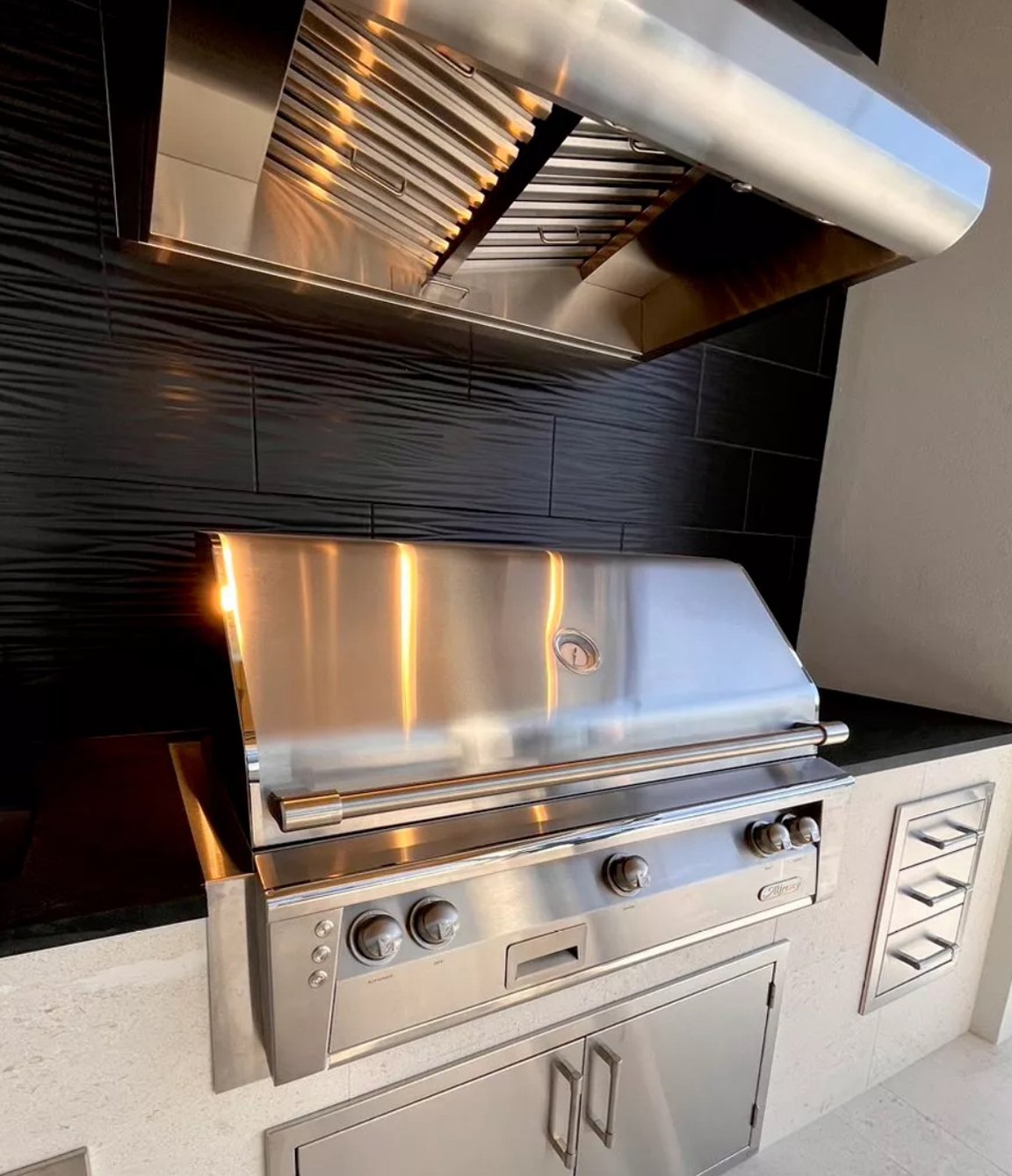 Stainless steel outdoor grill with control knobs, set on a light-colored countertop next to a black tile wall with textured pattern.