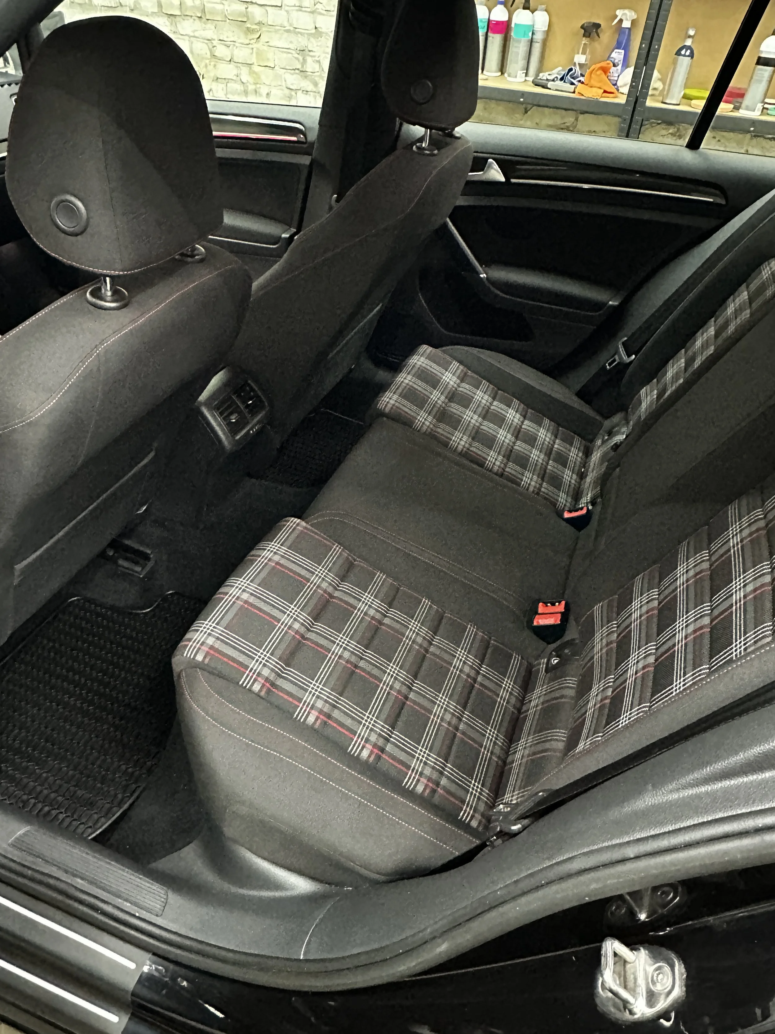 Das Bild zeigt die Rücksitze eines Autos mit einem karierten Sitzbezug, der auf zwei Sitzen angebracht ist. Im Hintergrund sind Regale mit Reinigungs- und Pflegeprodukten sichtbar.