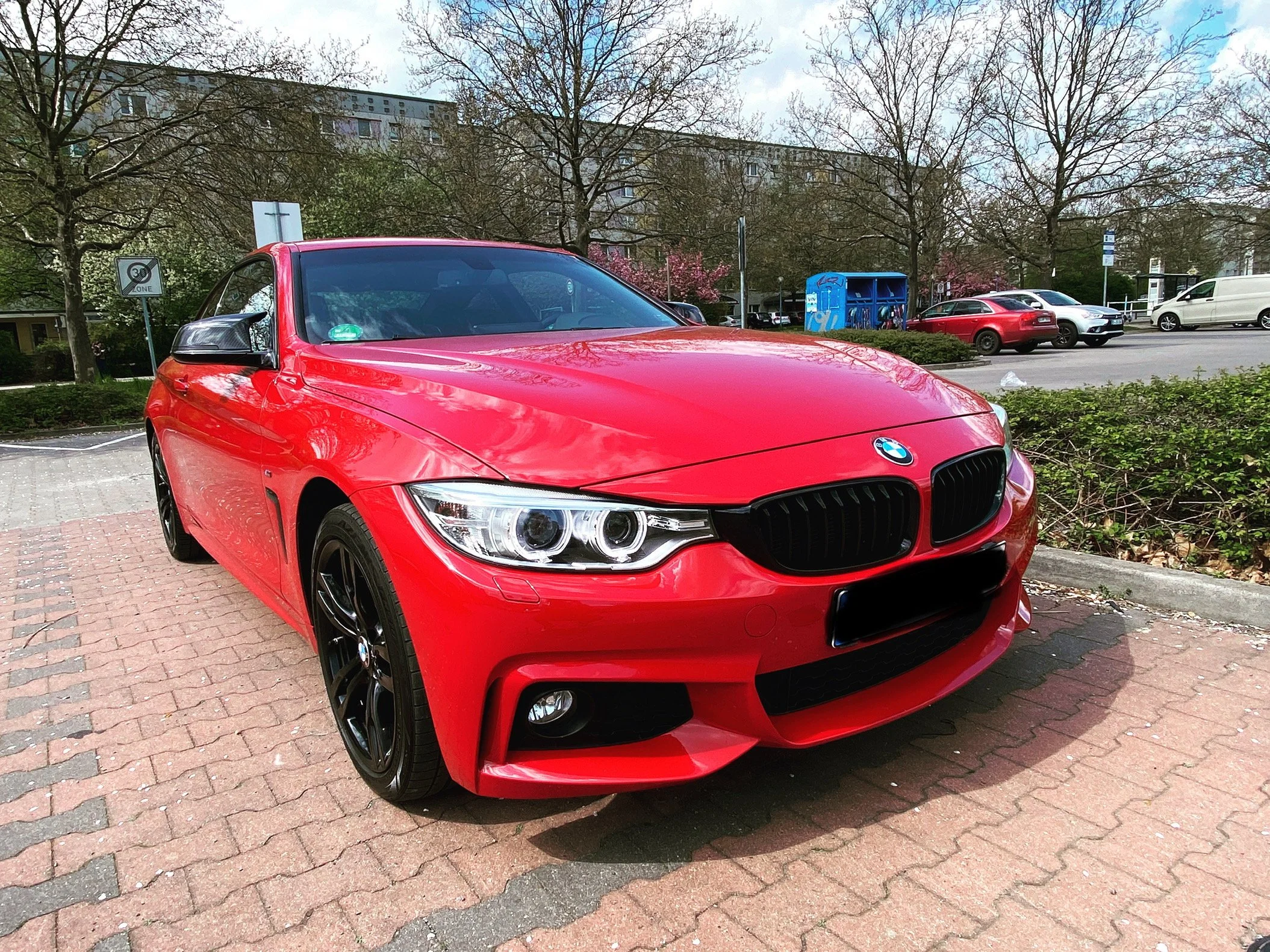 Roter BMW auf einem Parkplatz, umgeben von Bäumen und anderen Fahrzeugen, bei bewölktem Himmel.