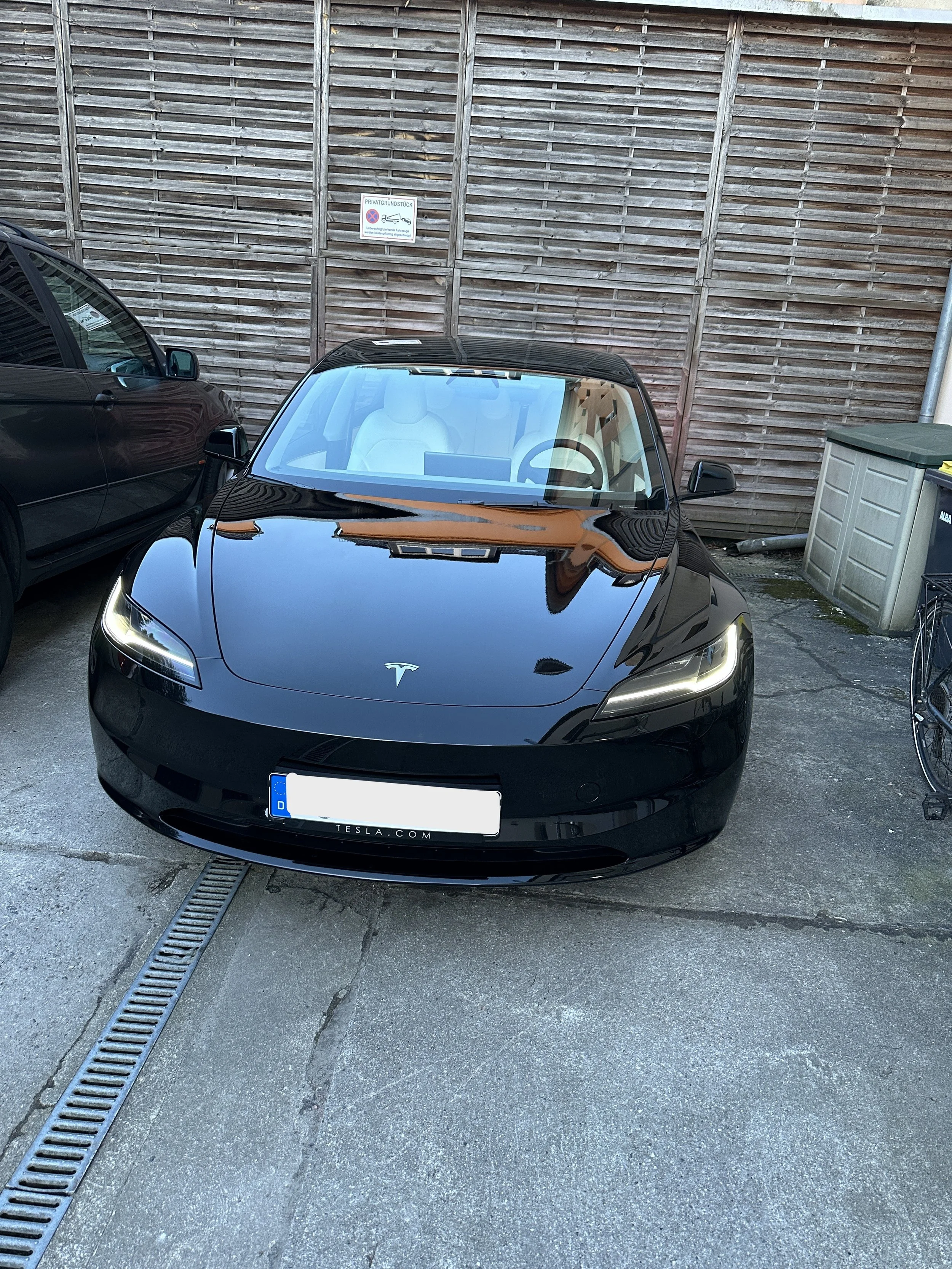 Ein schwarzer Tesla Roadster steht auf einem Parkplatz vor einem Holzzaun. Es ist ein Elektroauto, das von hinten und vorne sichtbar ist, mit einem niedlichen, modernen Design.