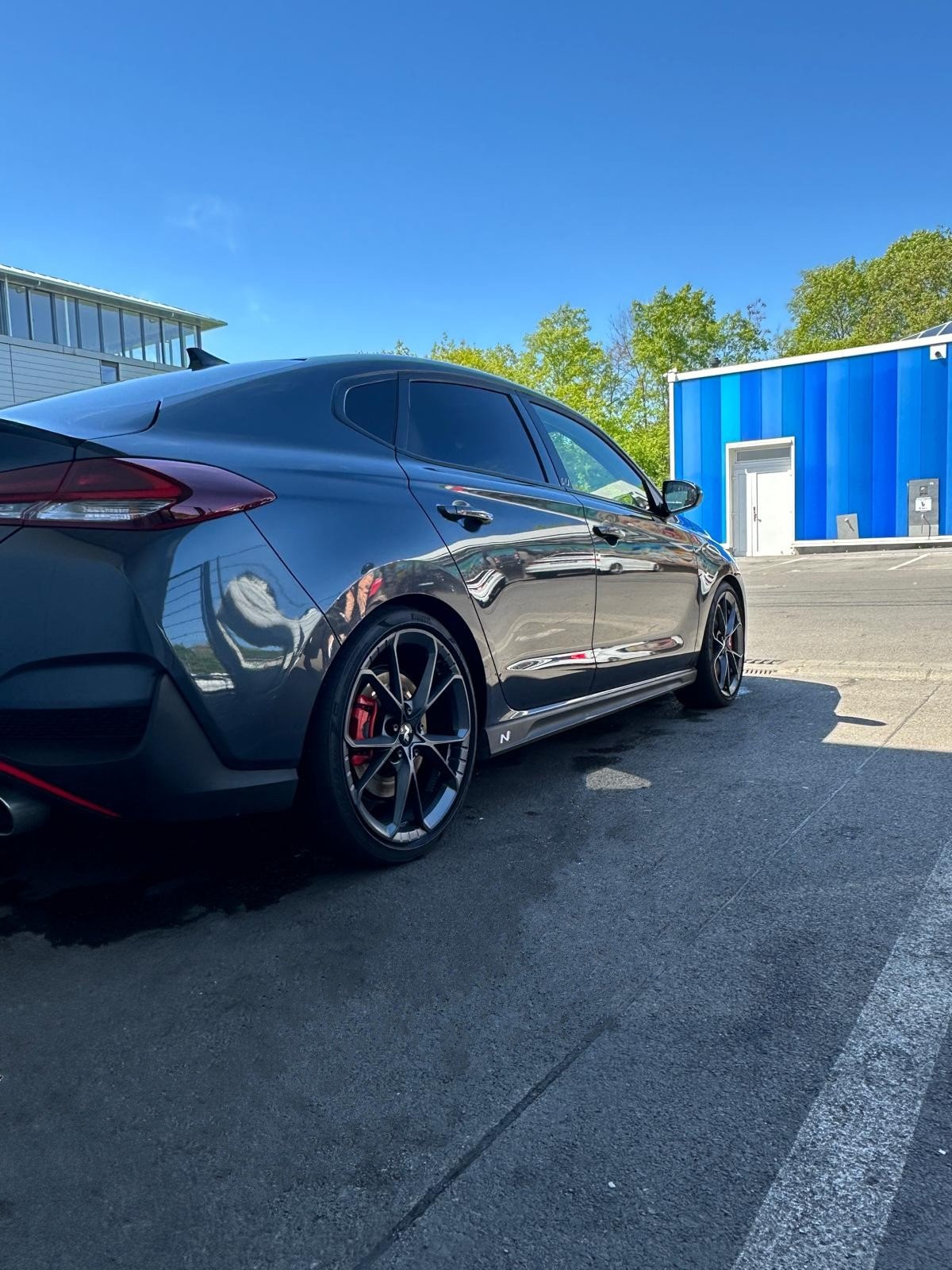 Ein schwarzes Auto, vermutlich ein Sportwagen, steht auf einem Parkplatz vor einem blauen Gebäude unter klarem Himmel, umgeben von Bäumen.