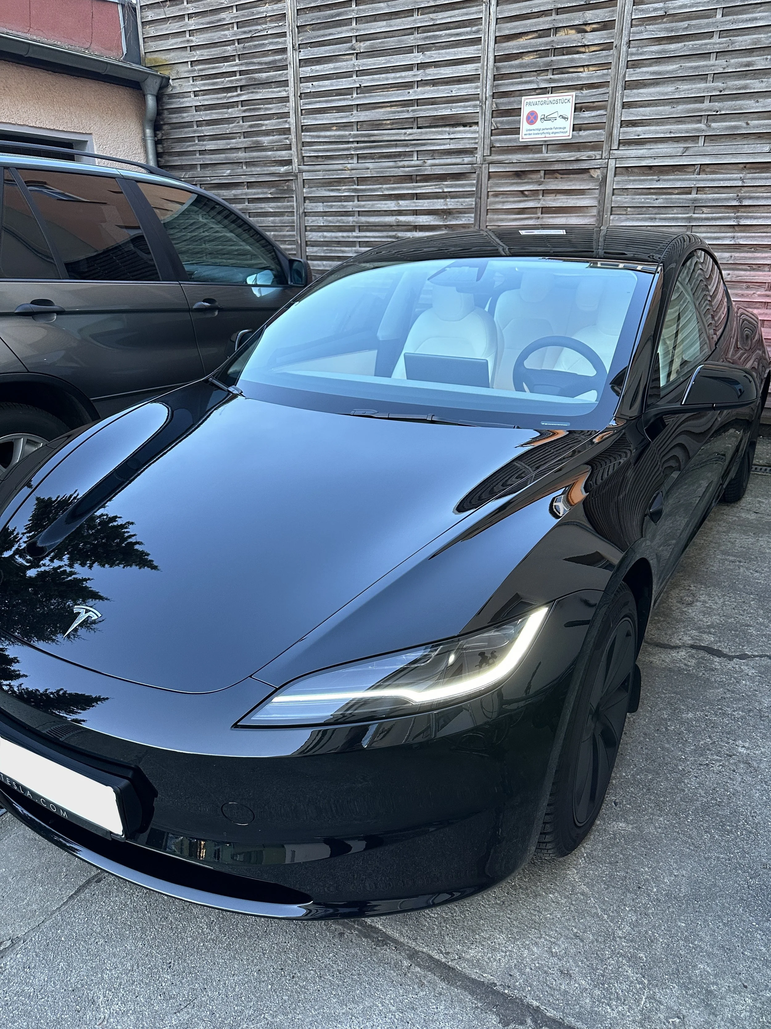 Ein schwarzer Tesla-Roadster parked auf einer Straße, neben einem weiteren schwarzen Fahrzeug, im Hintergrund eine Holzwand und ein privates Grundstücksschild.