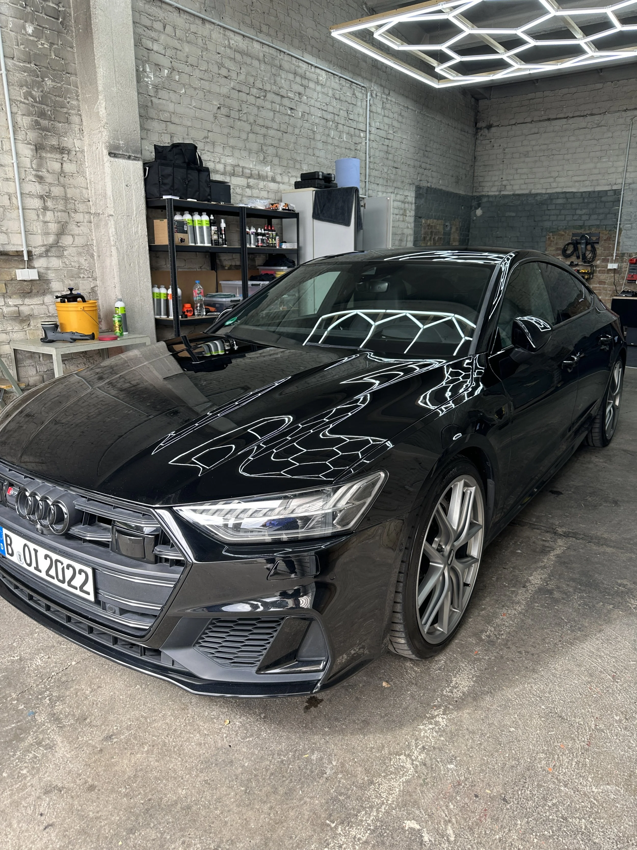 Ein schwarzer Audi Pkw in einer Garage, die Wände sind aus Bruchstein, im Hintergrund sind Werkzeuge und verschiedene Flaschen auf einem Regal sichtbar.
