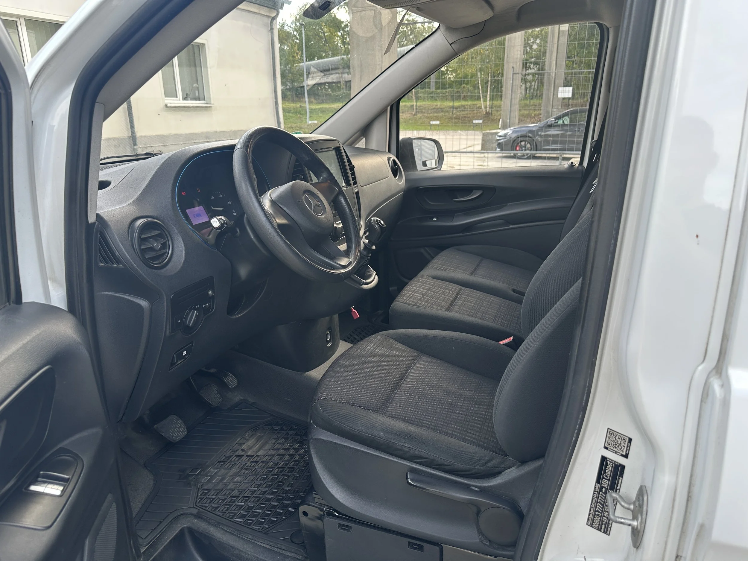 Der Innenraum eines Vans, mit schwarzem Armaturenbrett, Lenkrad und einer Sitzbank auf der Beifahrerseite, draußen sind einige Häuser und ein Auto zu sehen.