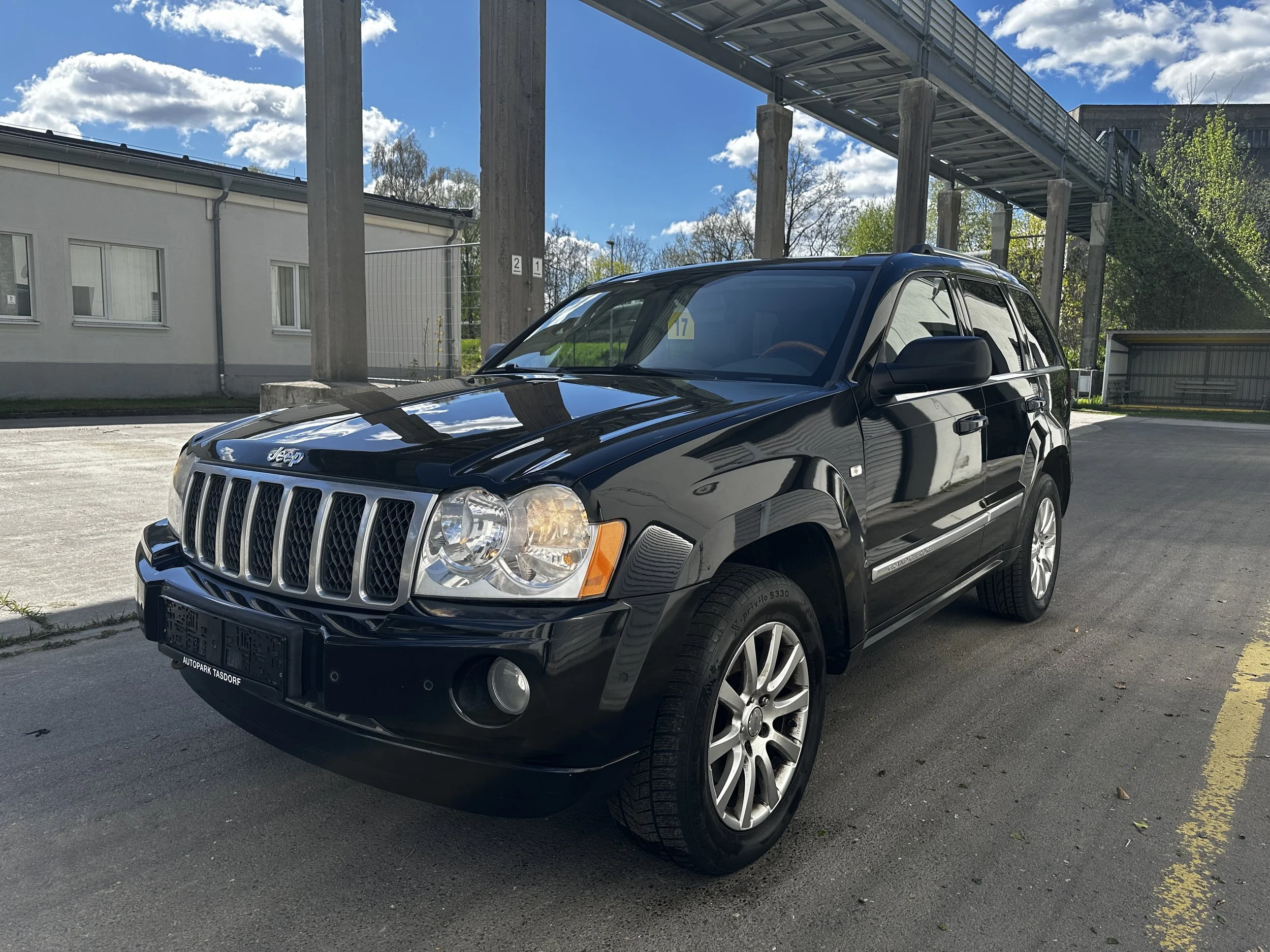 Schwarzer Jeep mit Silbern-Rädern im Parkhaus, im Hintergrund Himmel mit Wolken und ein überdachter Übergang mit Betonstützen.