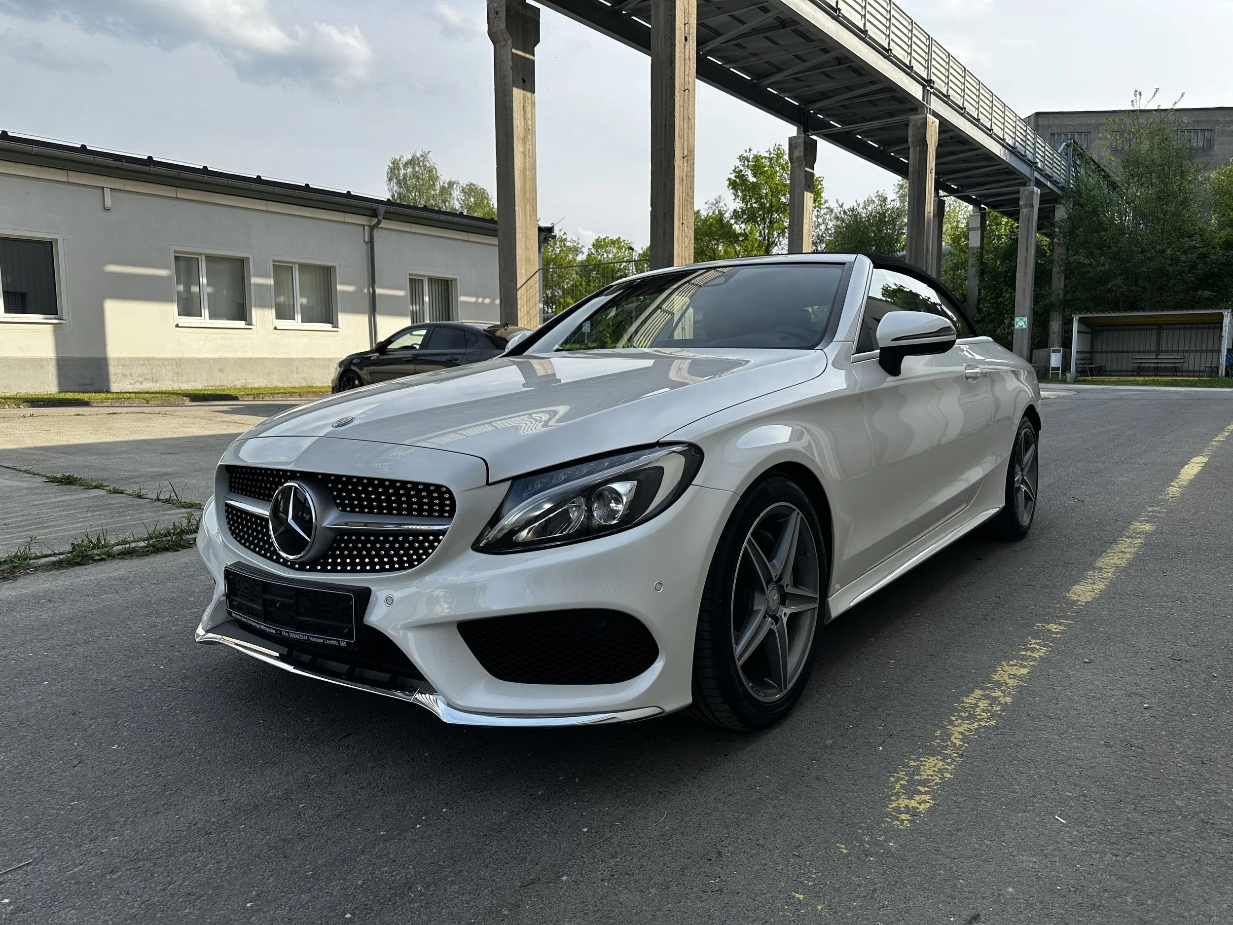 Ein weißer Mercedes-Benz Sportwagen parkt auf einem Parkplatz neben einem weißen Gebäude und einer Eisenbahnbrücke mit Betonpfeilern, umgeben von Bäumen.