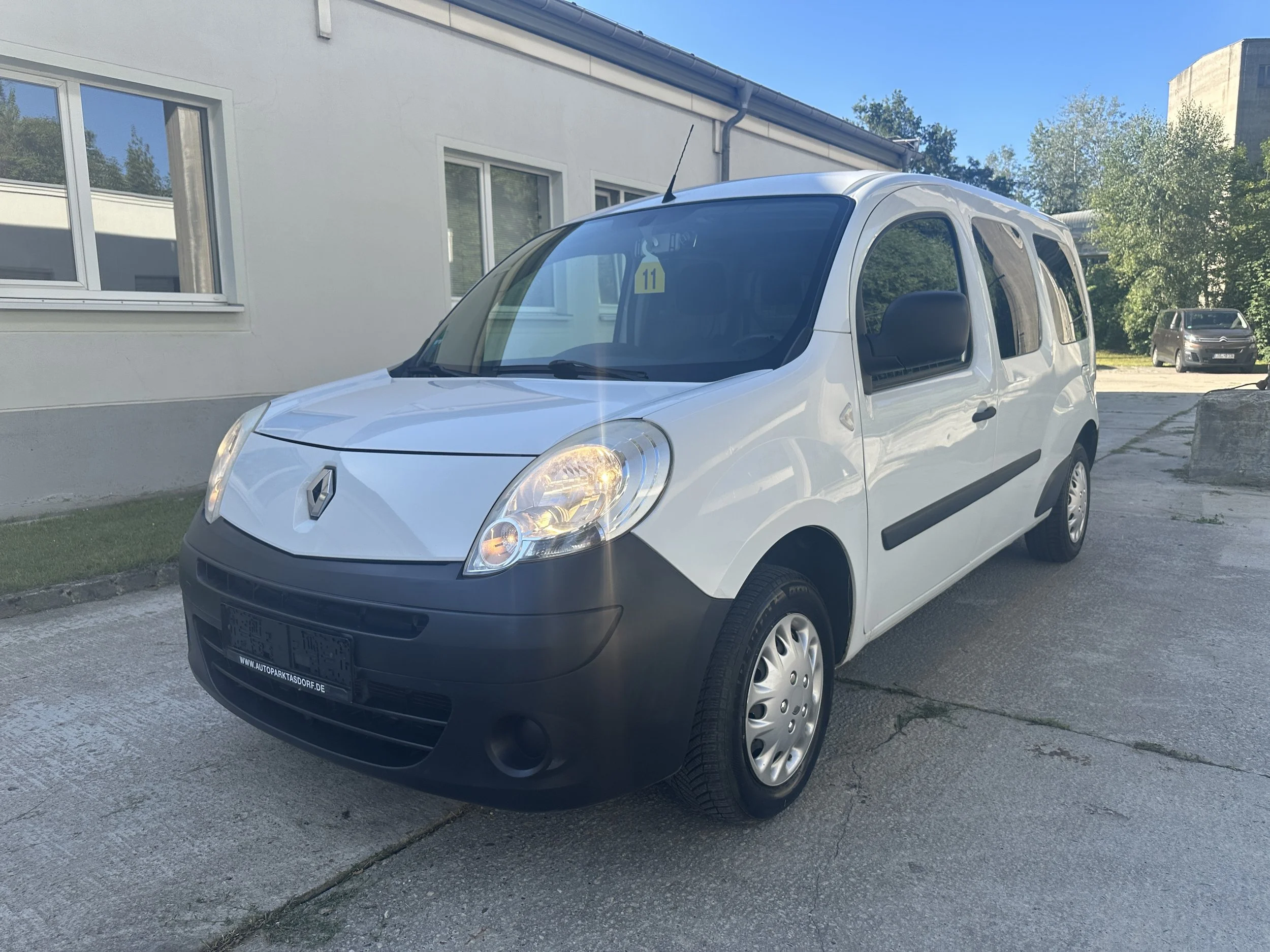 Weißer Renault Kangoo VW Touran auf Parkplatz vor einem weißen Gebäude, im Hintergrund Bäume und blauer Himmel.