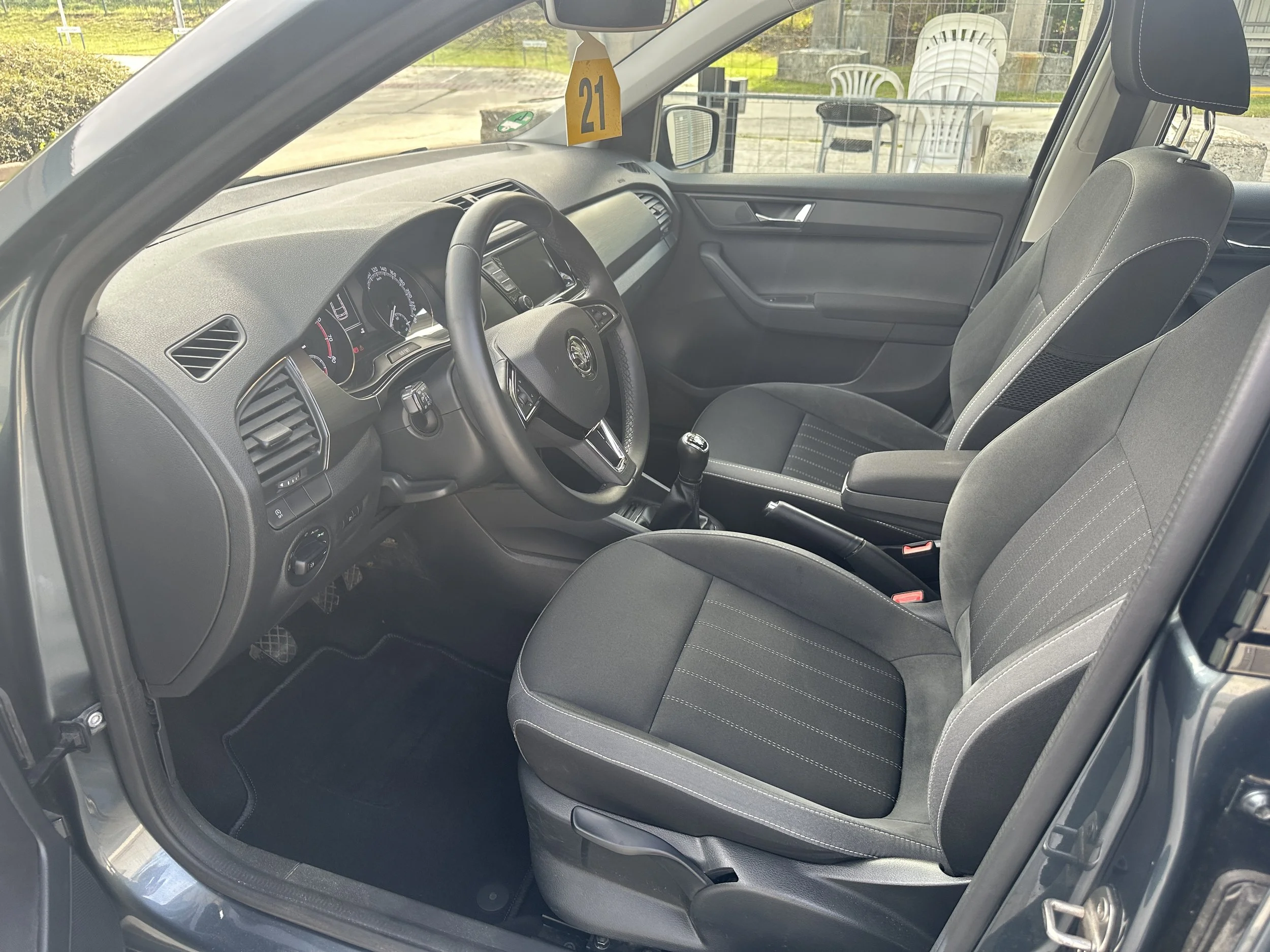 Innenansicht eines Autos mit schwarzem Sitz und Armlehne, schwarzem Lenkrad, manueller Schaltung, im Hintergrund eine Straße, eine Abbiegungsschilder und mehrere Stühle im Freien.