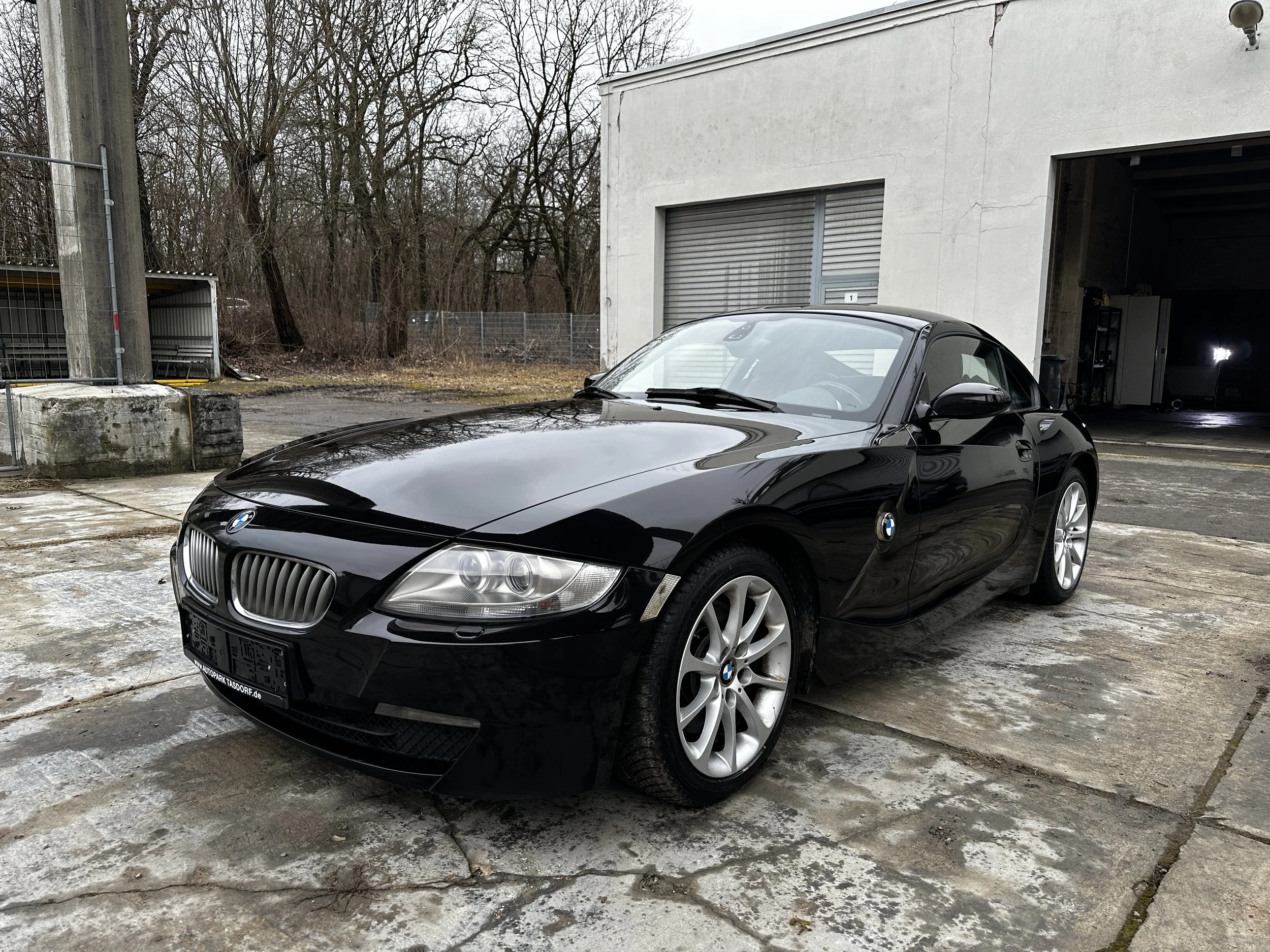Schwarzer BMW Z4 Cabrio vor einer Garage bei bewölktem Himmel, auf einer nassen Hoffläche mit Pfützen.