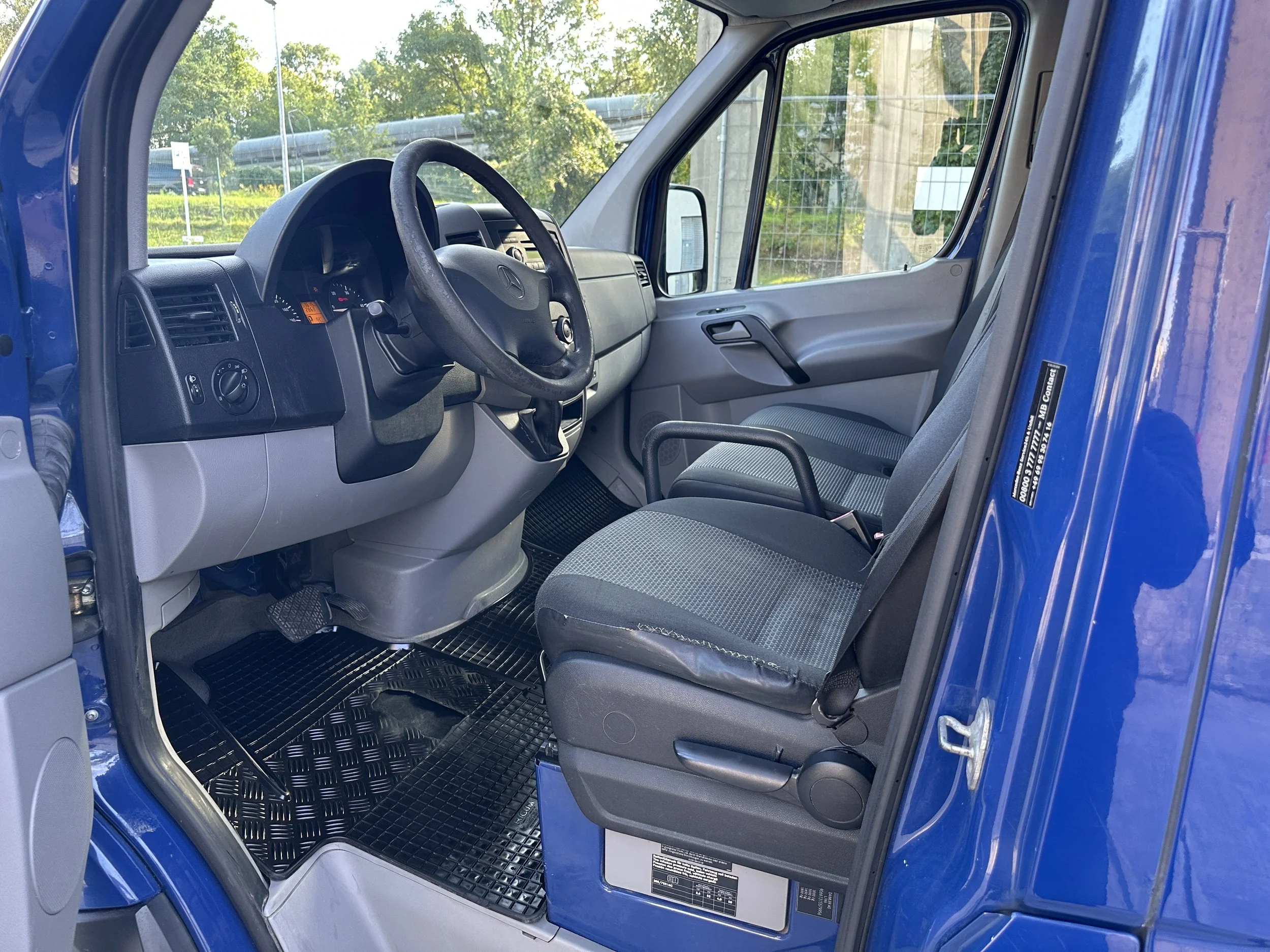 Innenansicht eines blauen Transporters, einschließlich Lenkrad, Fahrersitz, Beifahrersitz und Fenster mit Blick nach draußen, bei Tageslicht draußen mit Bäumen und einer Autobahn im Hintergrund.