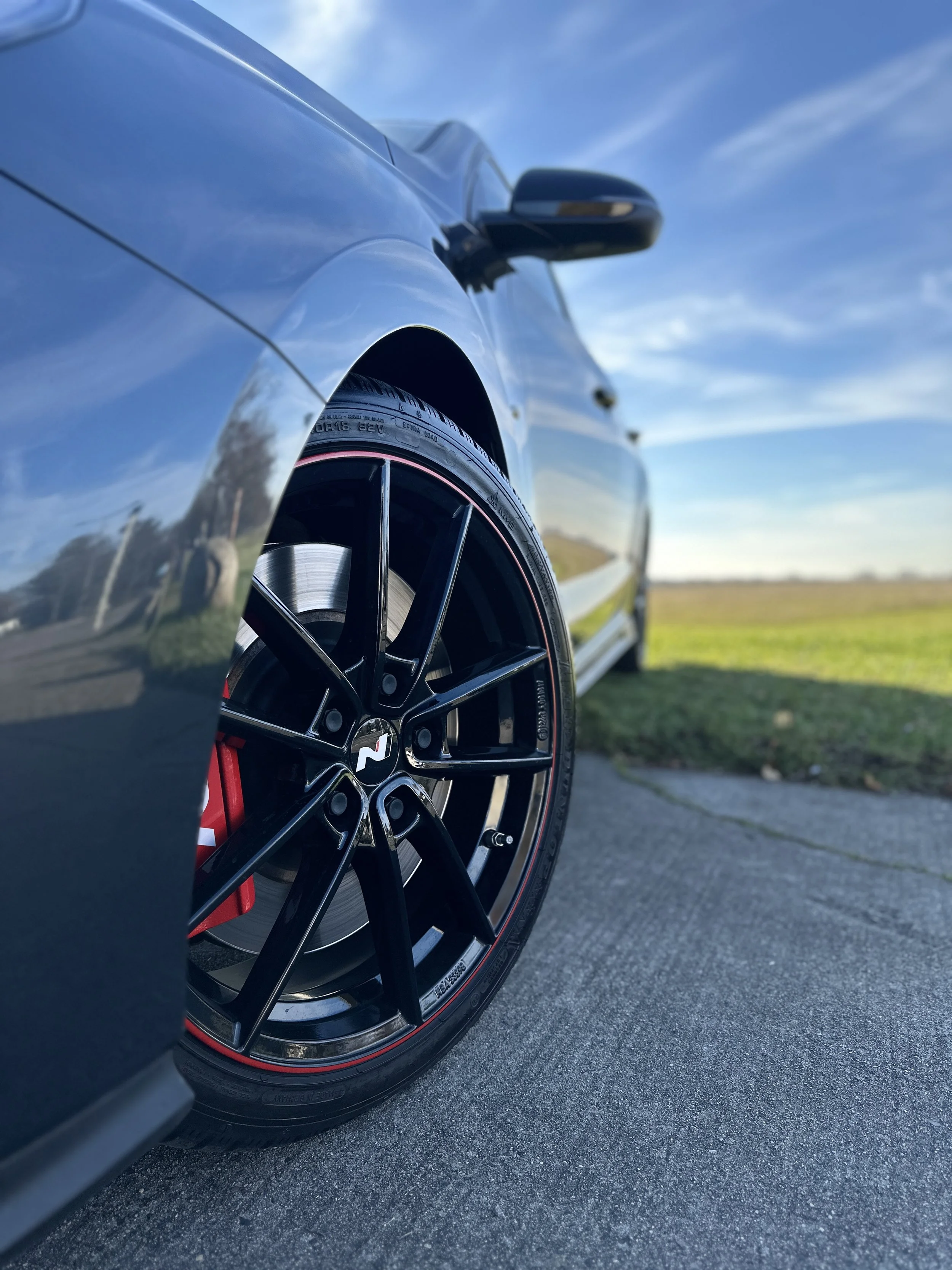 Nahaufnahme eines schwarzen Autos mit schwarzen Felgen und roten Bremssätteln, geparkt auf einer Straße mit grünem Feld im Hintergrund und blauem Himmel.