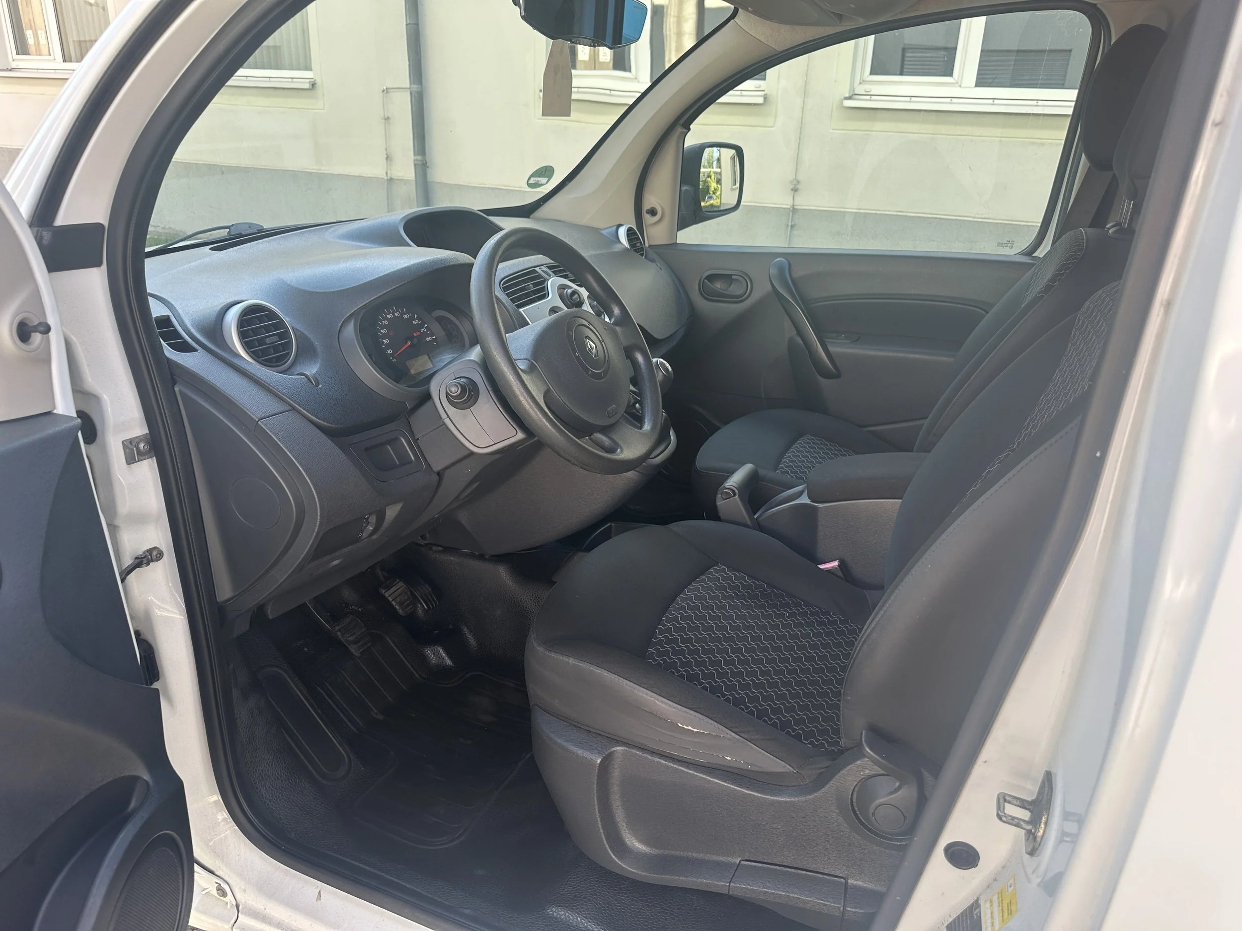 Der Innenraum eines Kleintransporters, sichtbar sind das Lenkrad, das Armaturenbrett mit Tachometer und Lüftungsöffnungen, sowie die Fahrermonitorseite ohne Fahrer. Die Sitze sind schwarz mit Muster, und das Fenster zeigt eine Außenwand mit Fenstern.