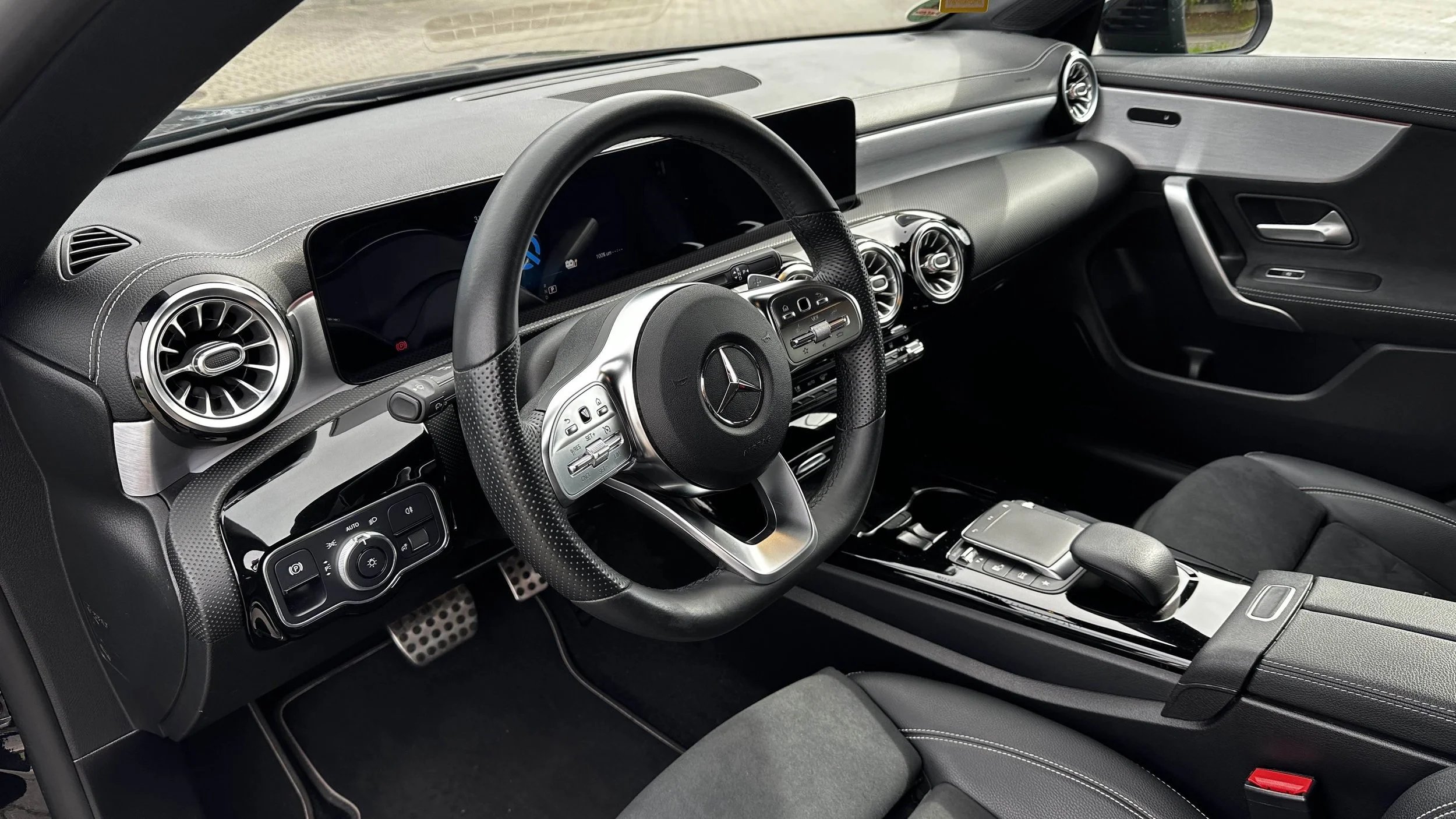 Interieur eines Mercedes-Benz Autos mit schwarzem Lenkrad, digitalen Armaturen, runden Luftauslässen, Mittelkonsole mit Touchpad, Schaltknauf und Armlehne.