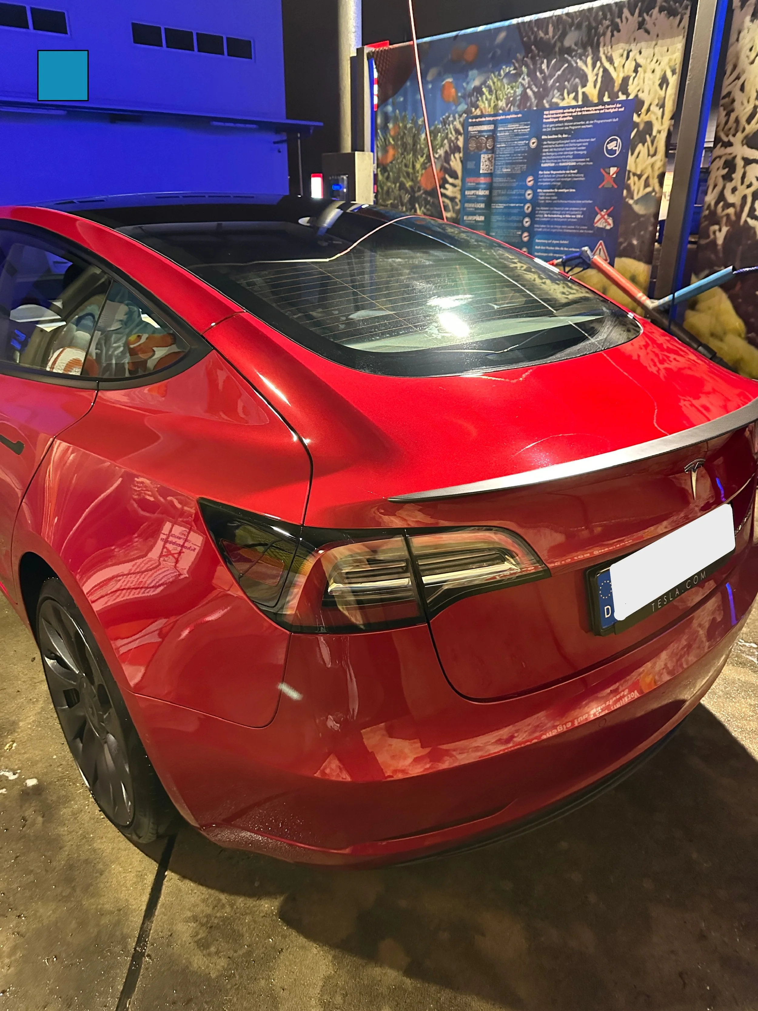 Rotes Elektroauto, vermutlich Tesla, an einer Ladestation. Der Hintergrund zeigt eine Informationstafel und eine Unterwasserwelt-Darstellung.