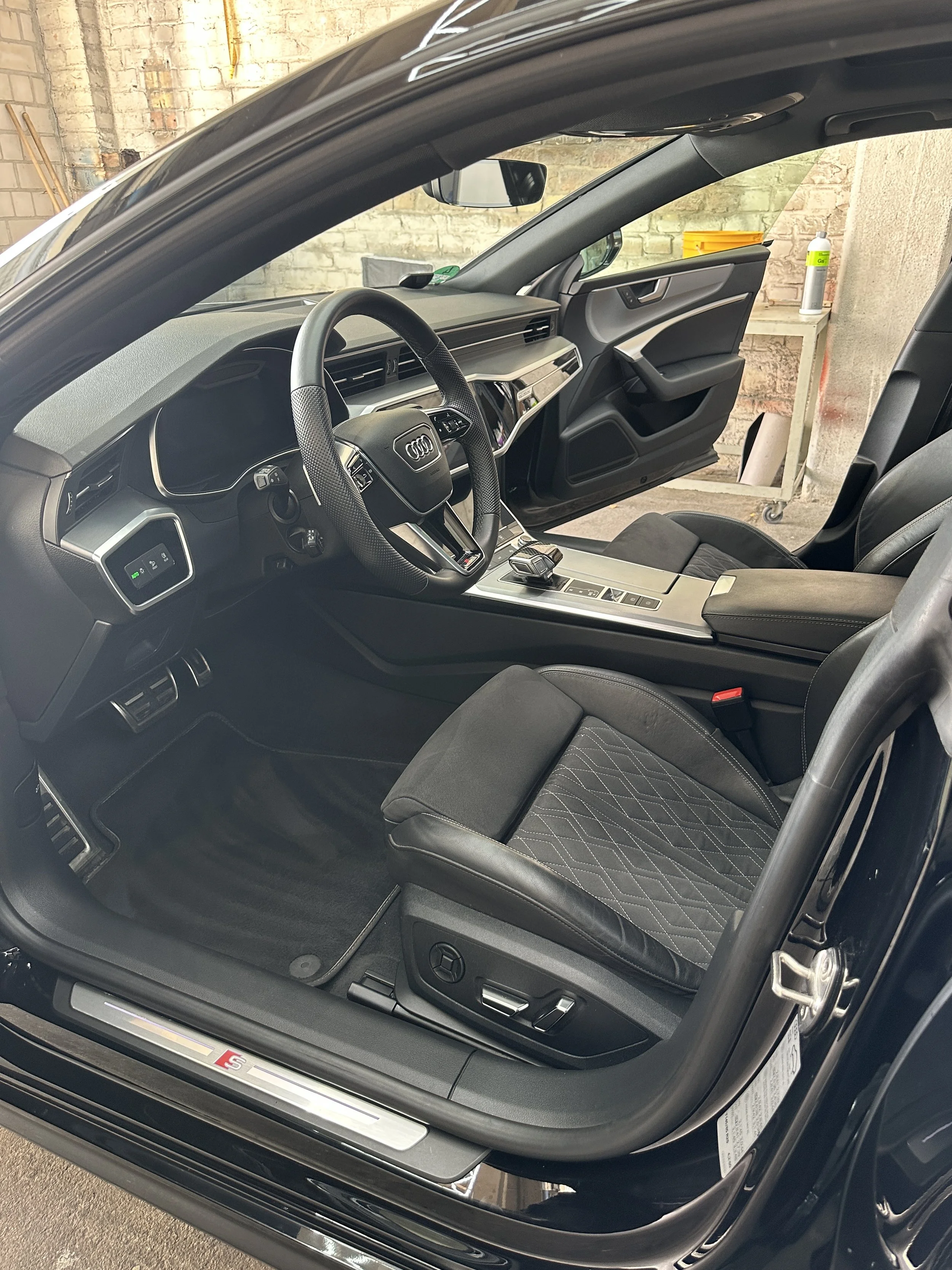 Der Innenraum eines schwarzen Autos, vermutlich ein Audi, mit Blick auf den Fahrersitz, das Lenkrad, die Mittelkonsole und die Tür. Im Hintergrund sieht man eine Werkstatt mit einem Metallwagen und Werkzeug.