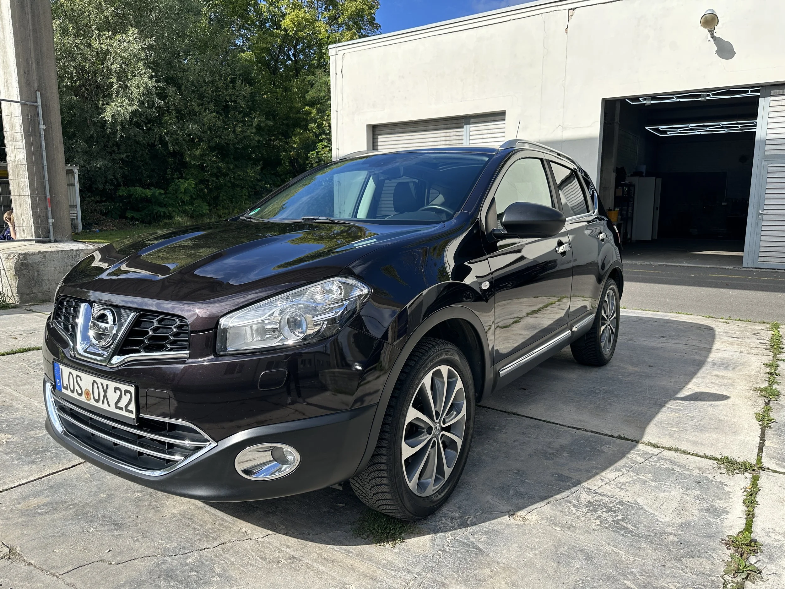 Ein schwarzer Nissan Juke SUV parkt vor einer weißen Garage mit offener Tür. Im Hintergrund sind Bäume und blauer Himmel sichtbar.
