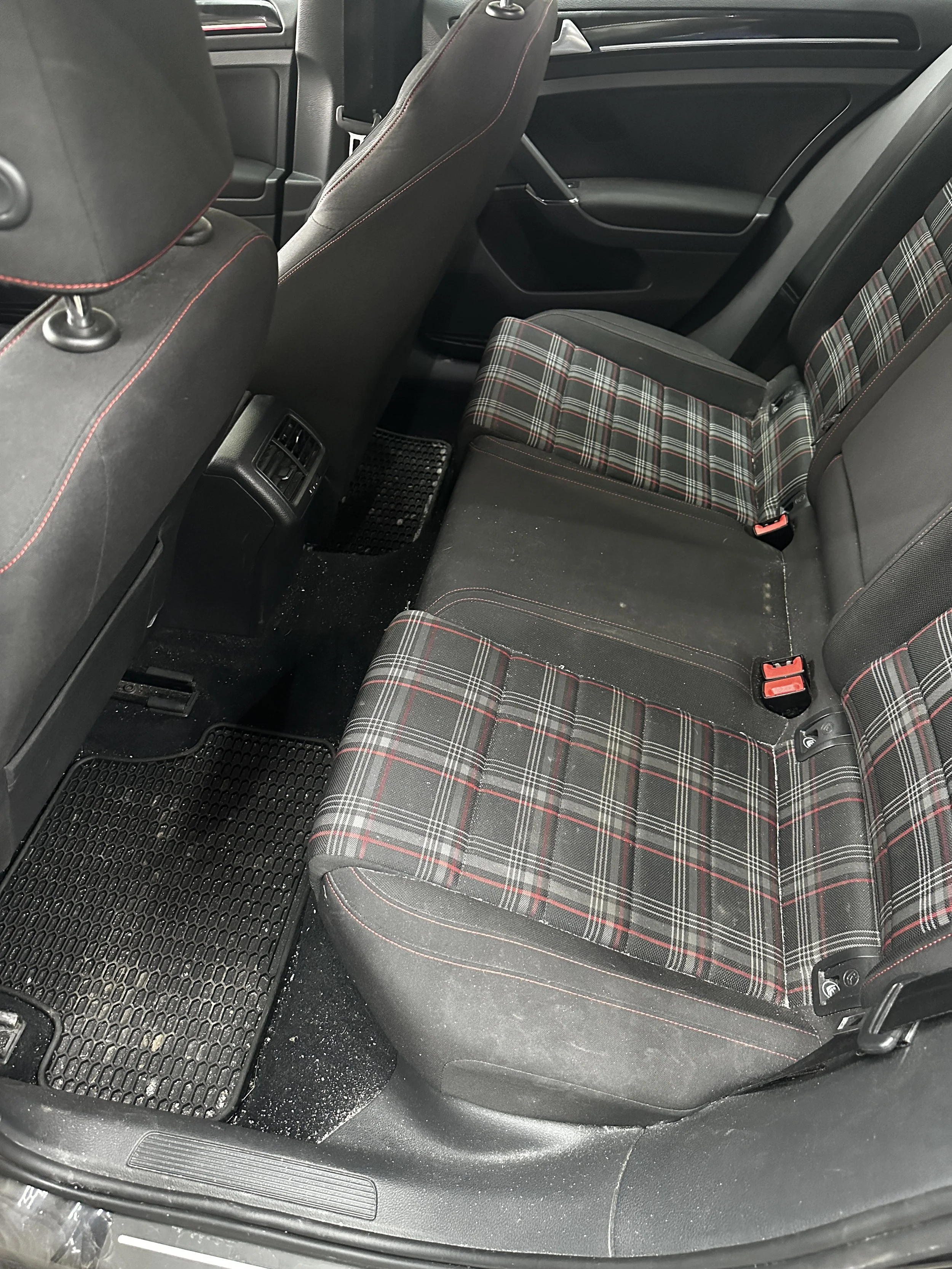 Der Innenraum eines Autos mit schwarzer Stoffpolsterung, rote Nähte und kariertes Muster auf den Sitzflächen. Es gibt Gittermatten auf dem Boden und Lüftungsöffnungen auf der Rückseite des Vordersitzes.