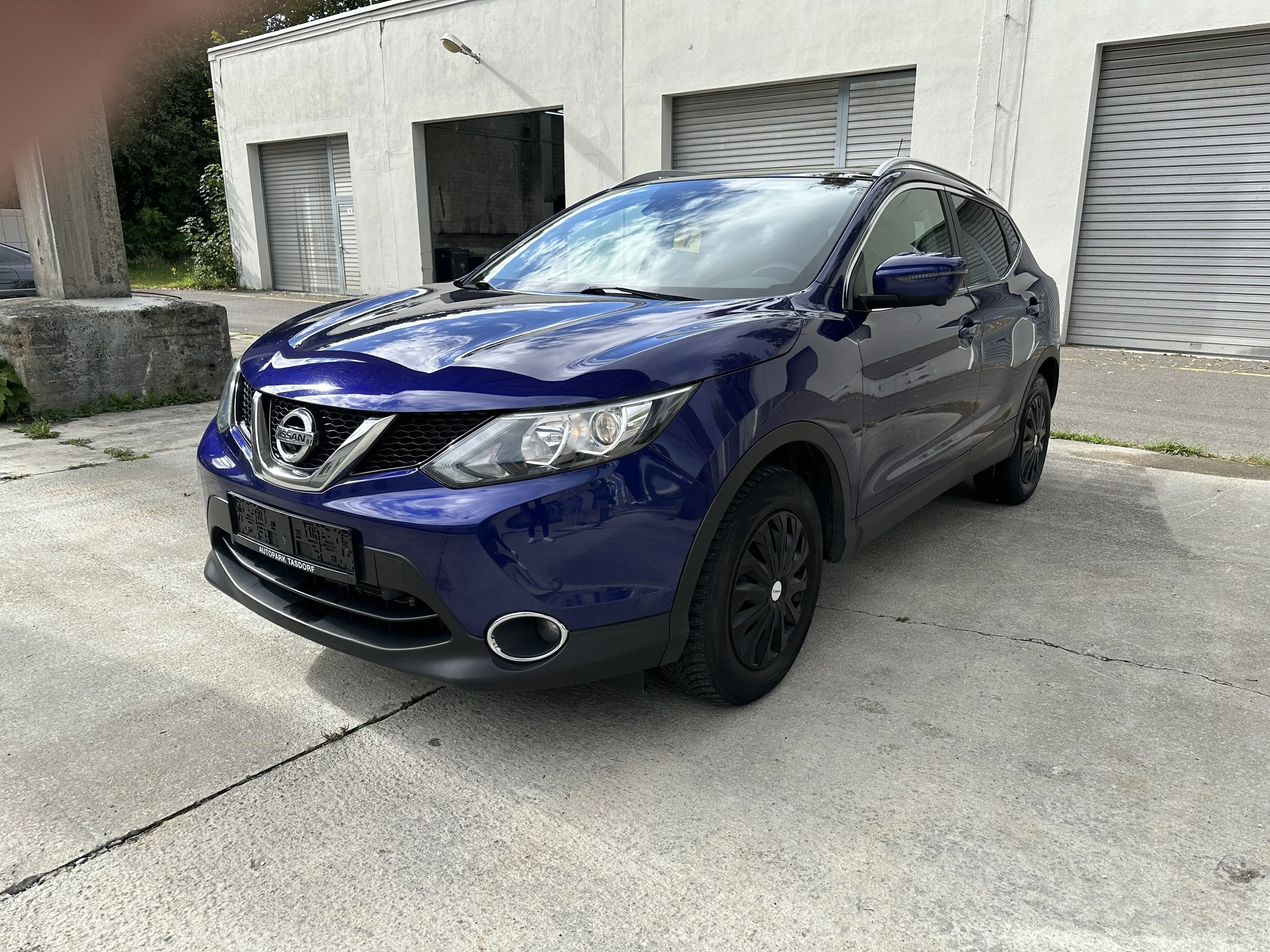 Ein blauer Nissan SUV ist vor einem grau gebauten Lagerhallen-Gebäude geparkt, im Hintergrund sind Baumzweige sichtbar.