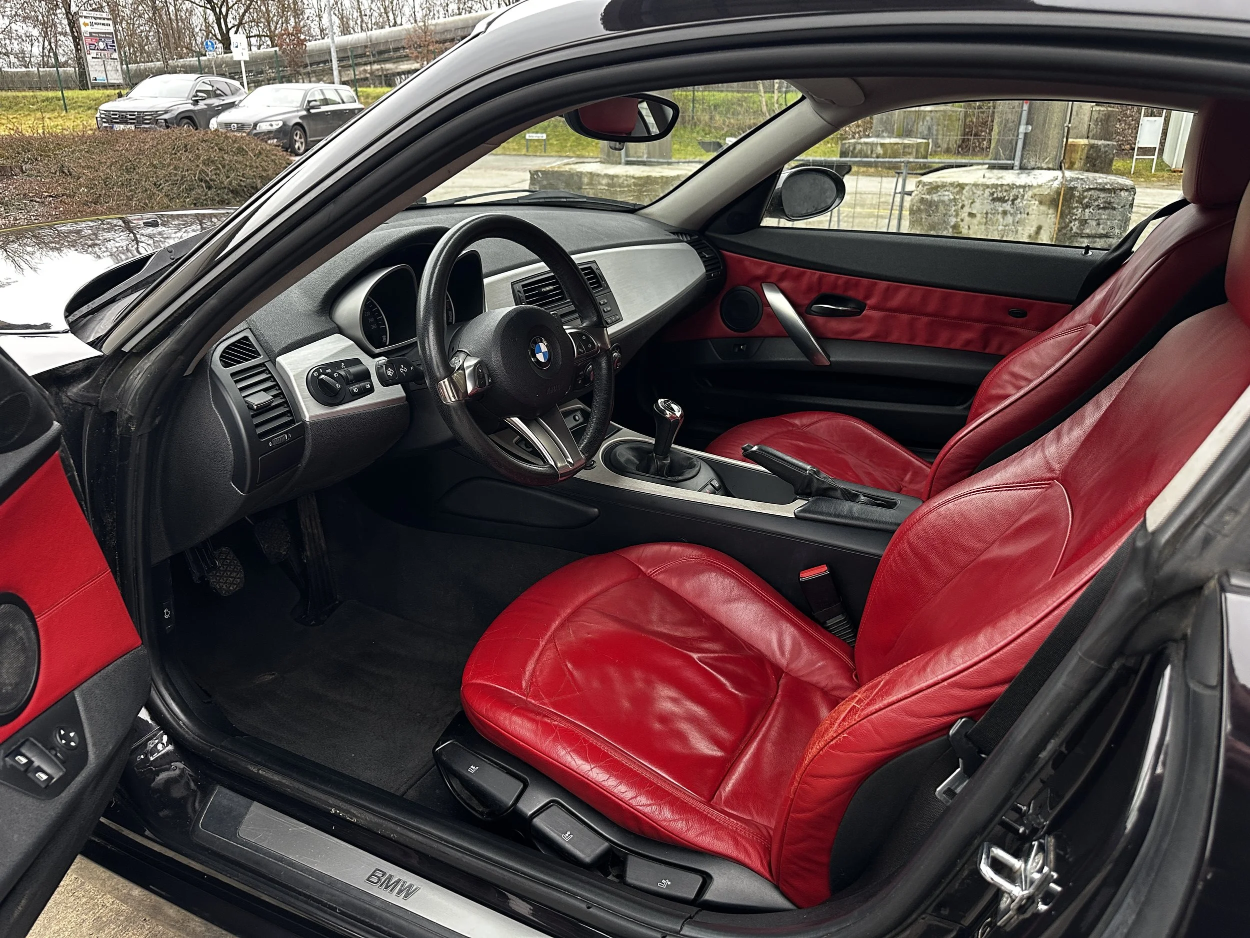 Innenansicht eines BMW Autos mit rotem Ledersitz, schwarzer Lenkrad, Mittelkonsole mit Schalthebel, Außensicht auf Parkplätze und Bäume durch die Fenster.