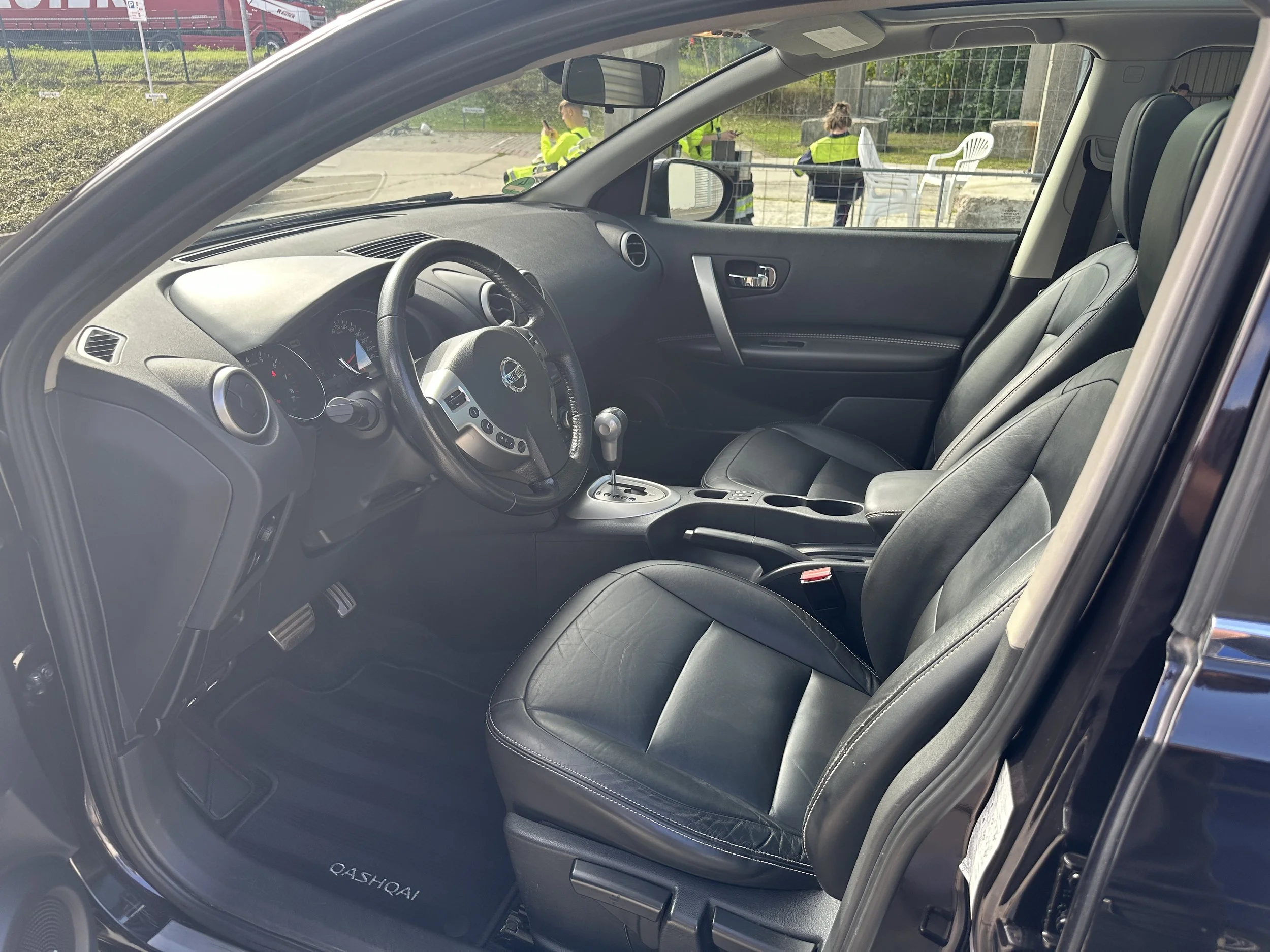 Der Blick ins Innere eines schwarzen Autos mit Ledersitzen, including das Lenkrad, das Armaturenbrett und die Sitze. Im Hintergrund sind Personen in leuchtend gelben Sicherheitswesten, die draußen stehen und sich unterhalten.