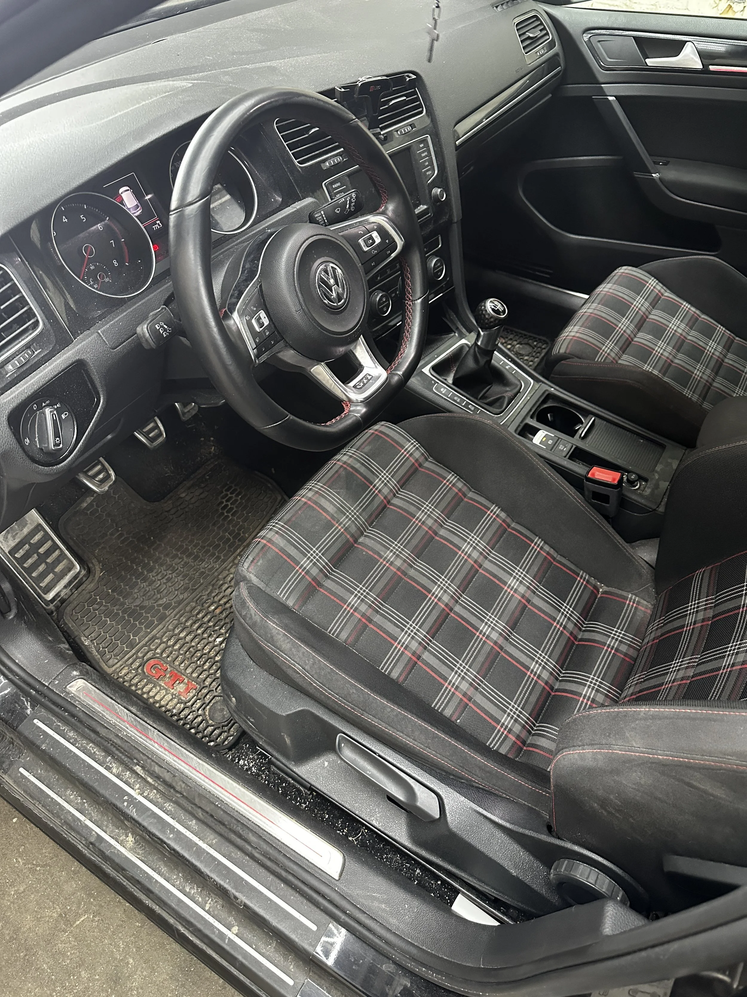 Innenraum eines Volkswagen Golf GTI, schwarz mit grauer und roter Karosserie, mit Schaltknüppel, Sportsitzen mit Tartanmuster, Pedale, Armaturenbrett, Lenkrad und Infotainment-System sichtbar.