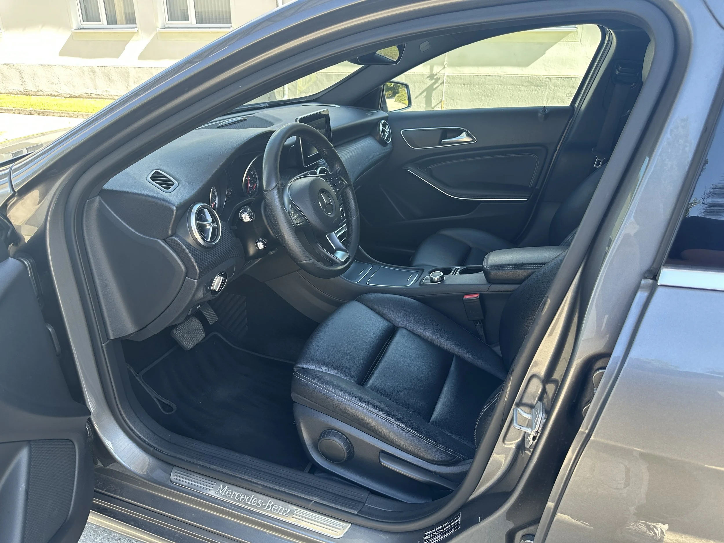 Innenraum eines schwarzen Mercedes-Benz Autos, Blick auf das Lenkrad, die Armaturen und den Sitz auf der Fahrerseite. Es ist hell und modern gestaltet.
