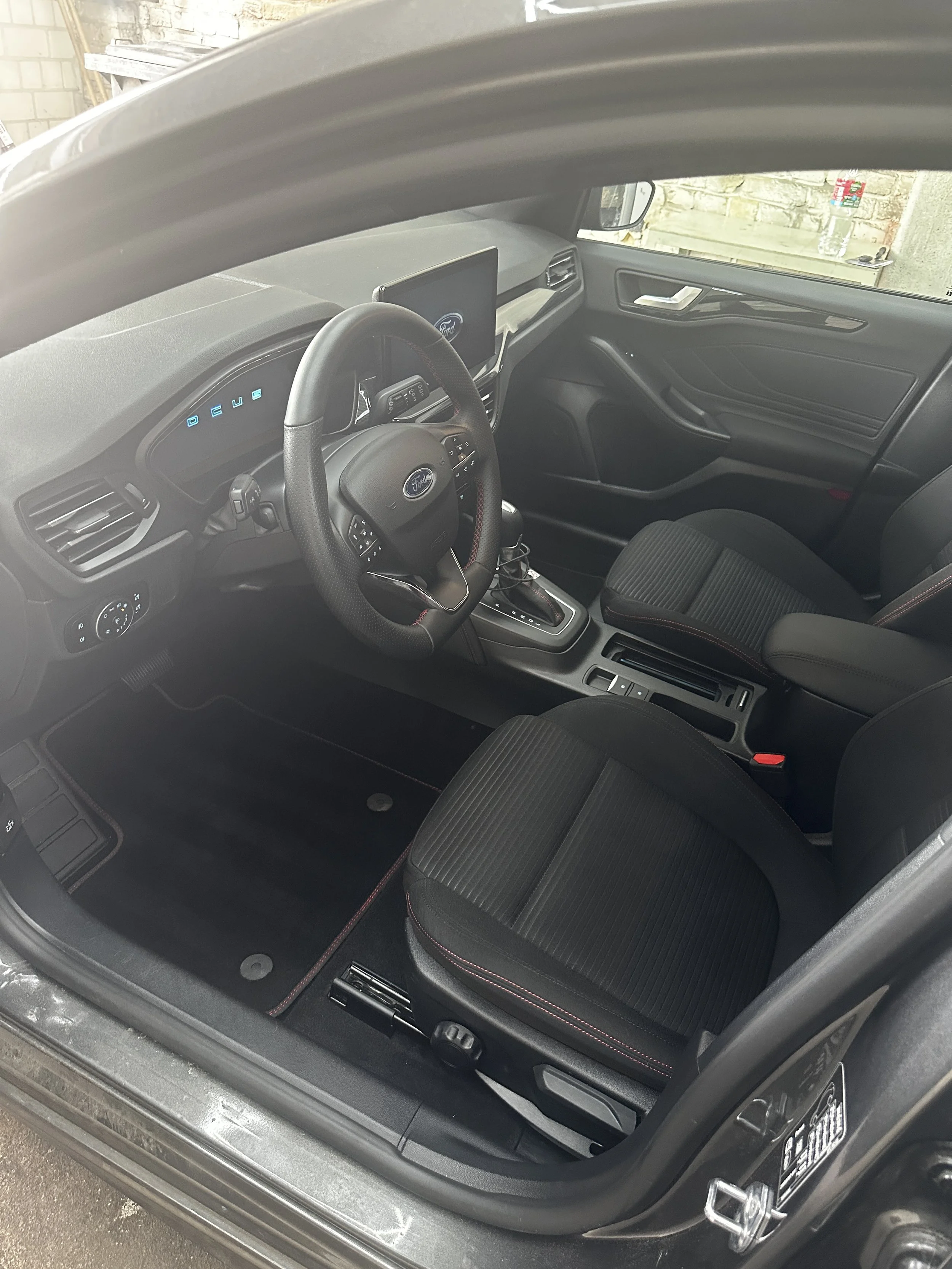 Innenraum eines Autos, Blick auf das Lenkrad, digitaler Tachometer, Infotainmentsystem, Schalthebel, Fahrersitz und Tür mit Griff.