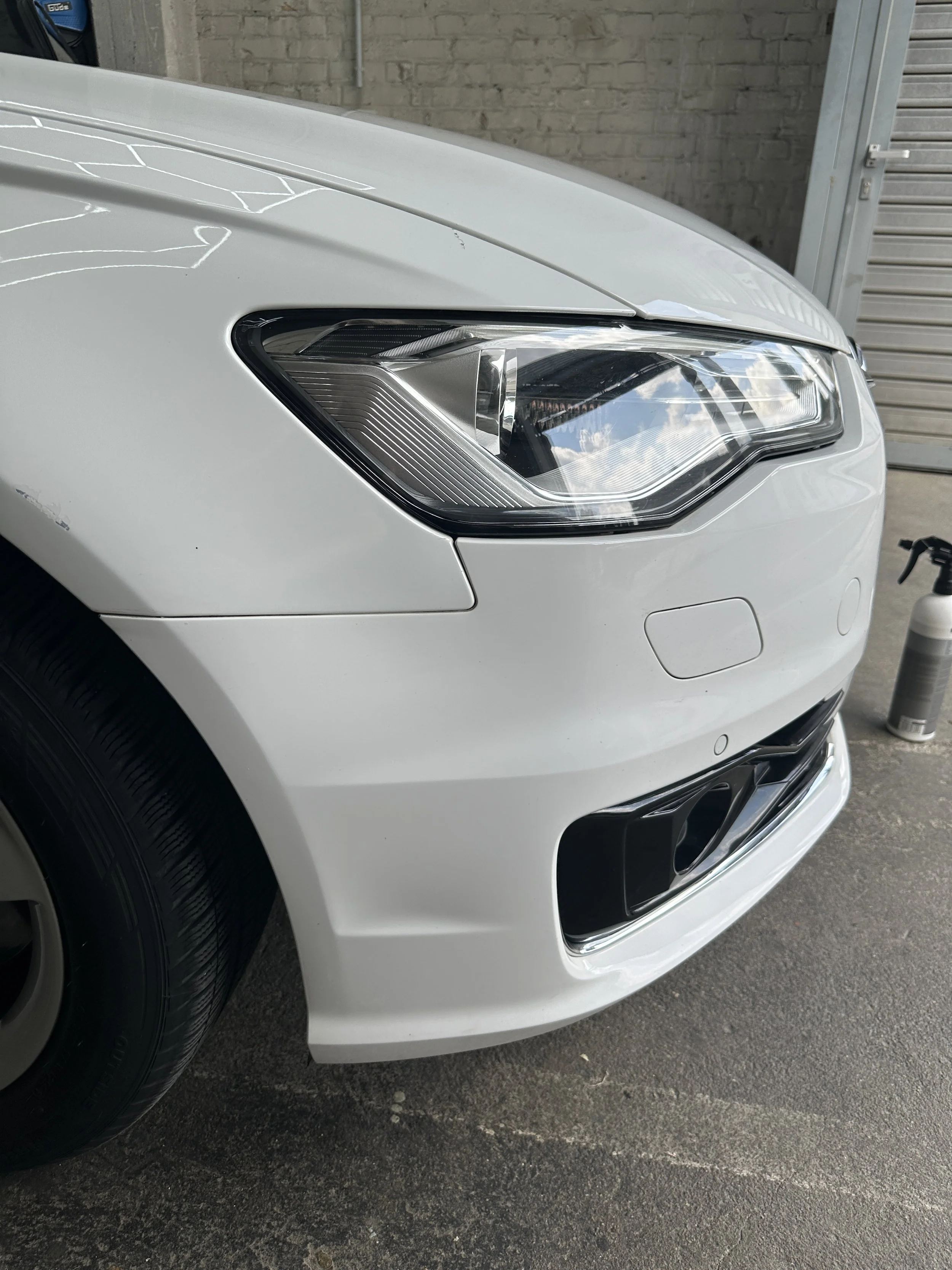 Vorderseite eines weißen Autos, Blick auf die Scheinwerfer und Stoßstange, im Hintergrund ein graues Backsteinmauer, Teil eines Garagentores und ein Ölkanister.