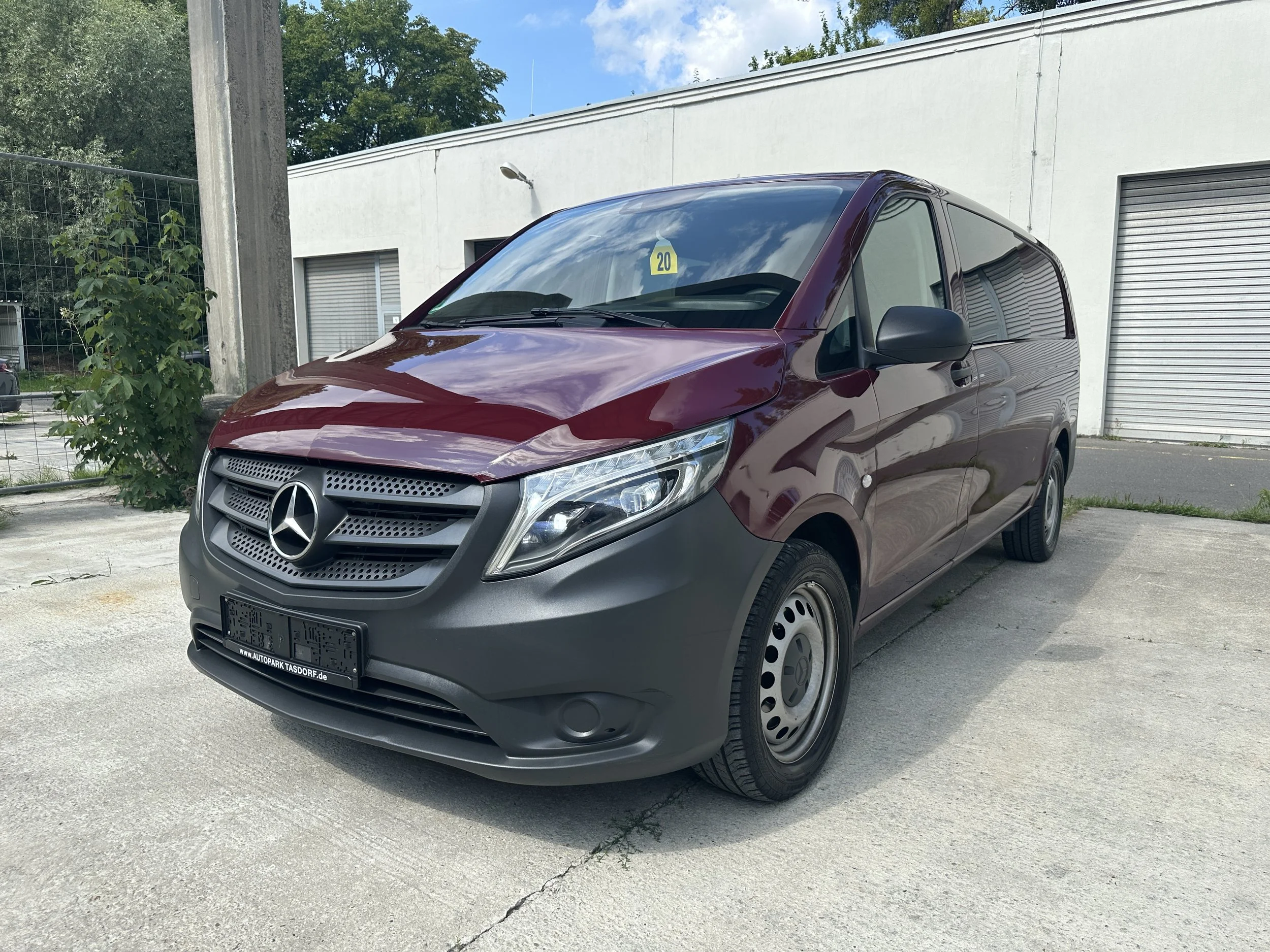 Roter Mercedes-Benz Transporter vor einer weißen Wand mit Rollladen, parkend auf einer betonierten Fläche, neben einer kleinen Pflanze und einem Zaun im Hintergrund.