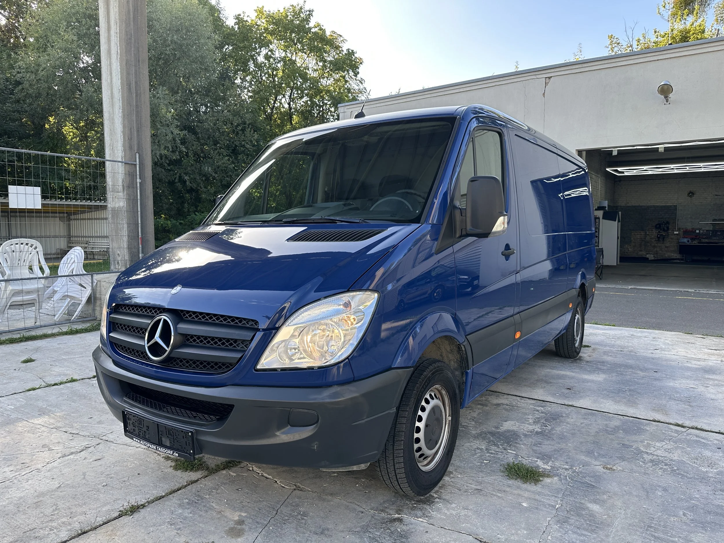 Ein blauer Mercedes-Benz Transporter parkt auf einem Parkplatz vor einer Garage. Im Hintergrund sind Bäume, ein Strommast, weiße Stühle und eine weiße Wand zu sehen.