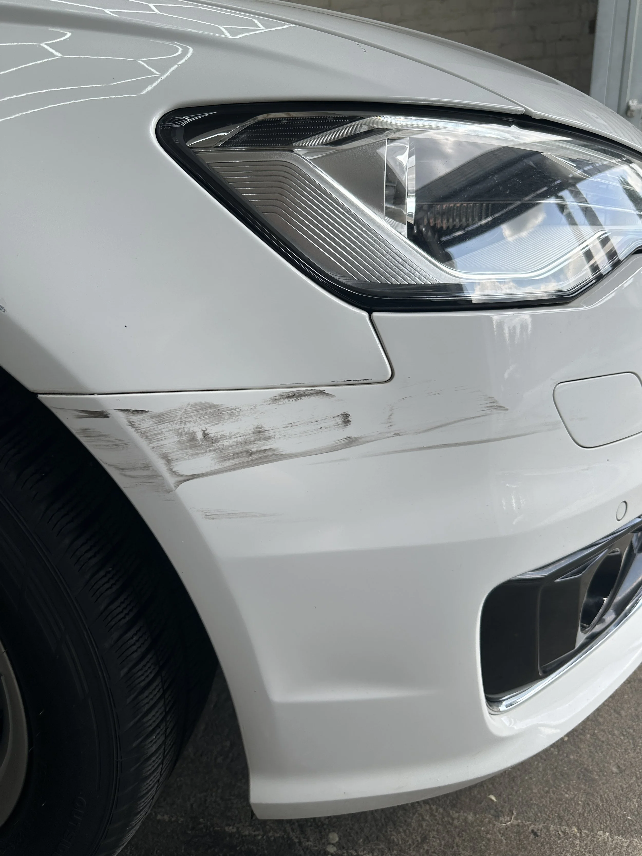 Nahaufnahme der Autoserie, Fokus auf die Scheinwerfer und vordere Stoßstange eines weißen Autos, sichtbarer Reifen und Teil des Kotflügels.