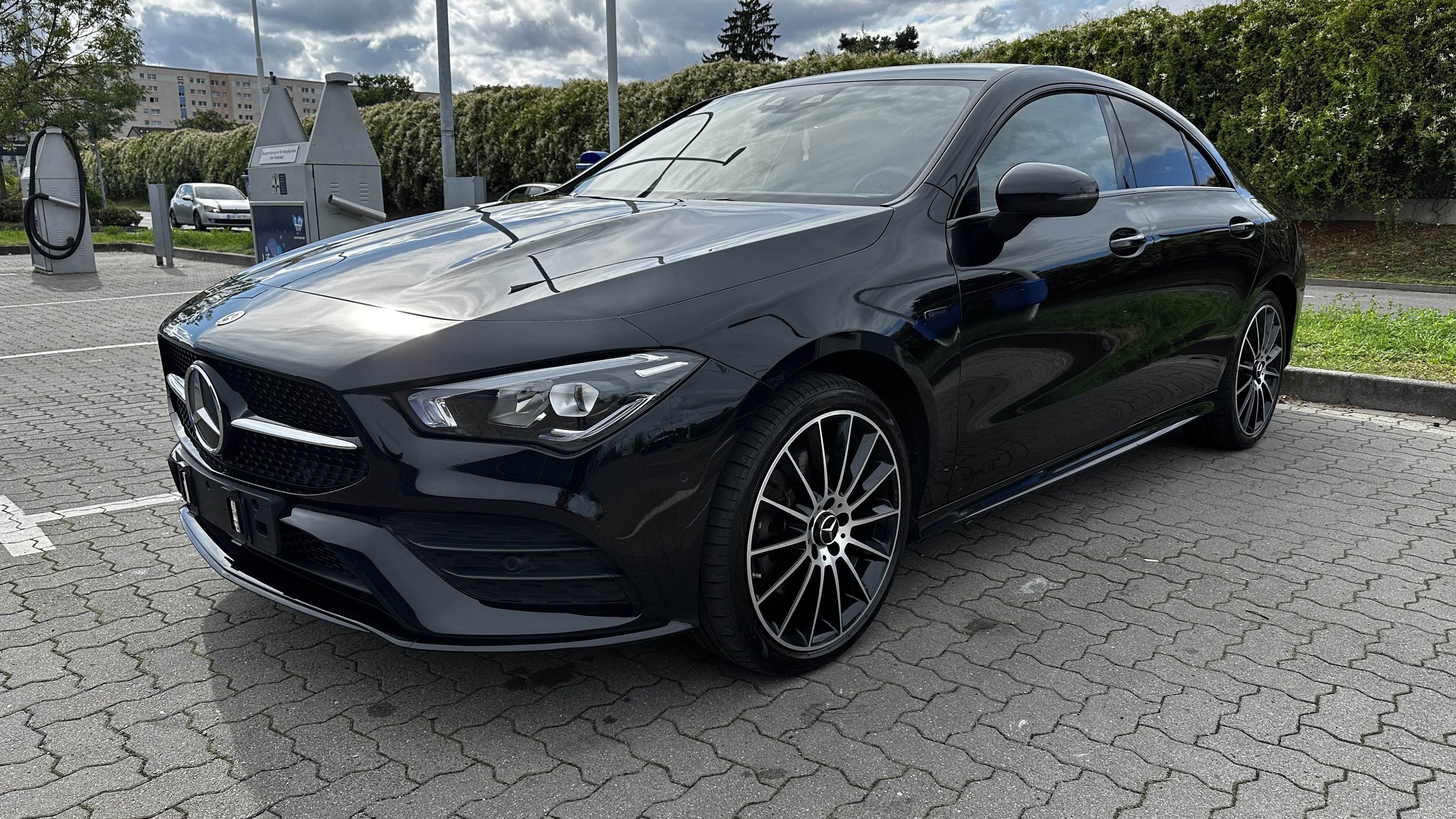 Schwarzer Mercedes-Benz Limousine parkt auf einem Parkplatz mit Pflastersteinen, im Hintergrund Büschen, eine Person und eine Hausreihe.