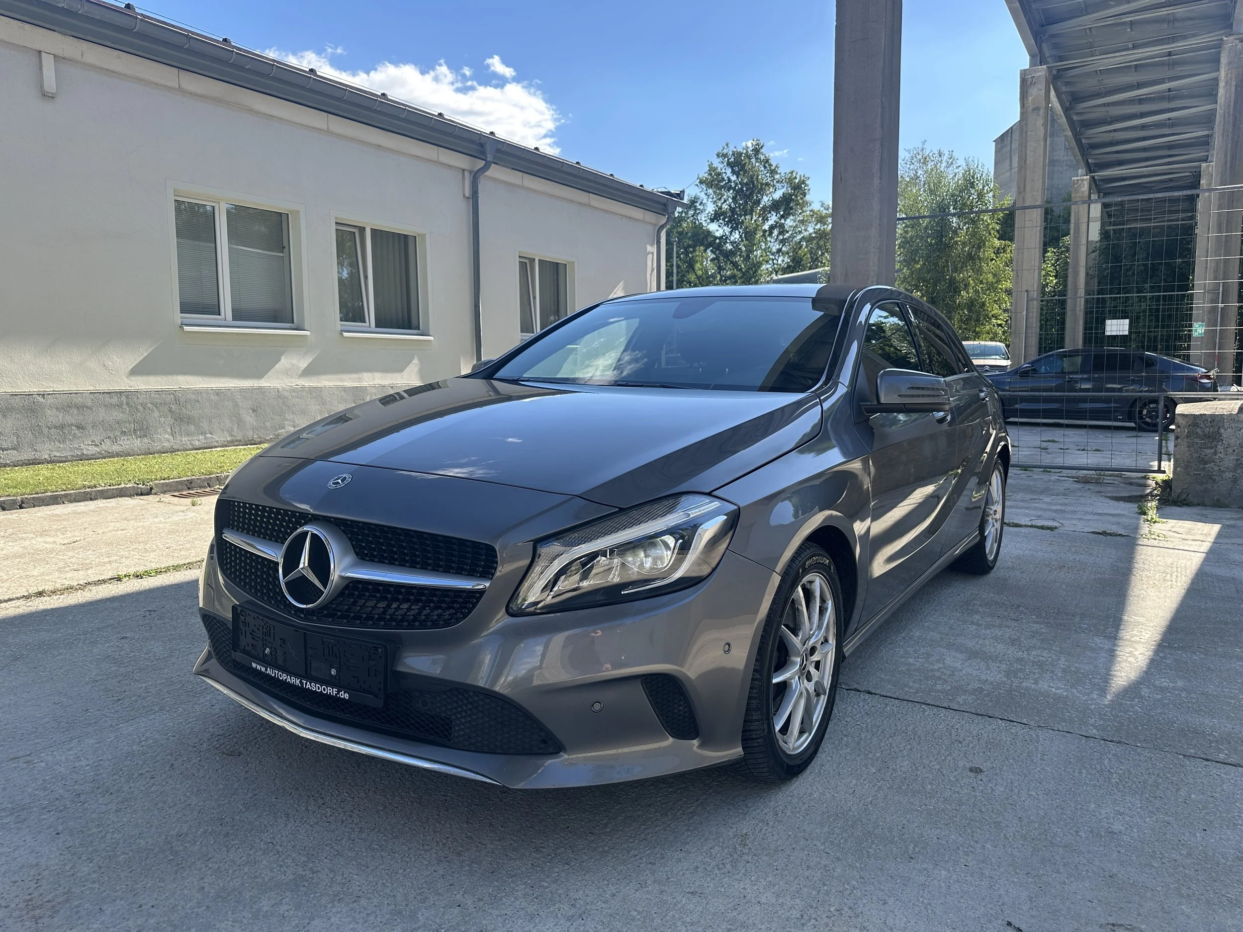 Schwarzer Mercedes-Benz-Auto vor einer weißen Hauswand, stehend auf Asphalt, bei sonnigem Himmel mit Wolken, im Hintergrund Baugerüst und andere Fahrzeuge.
