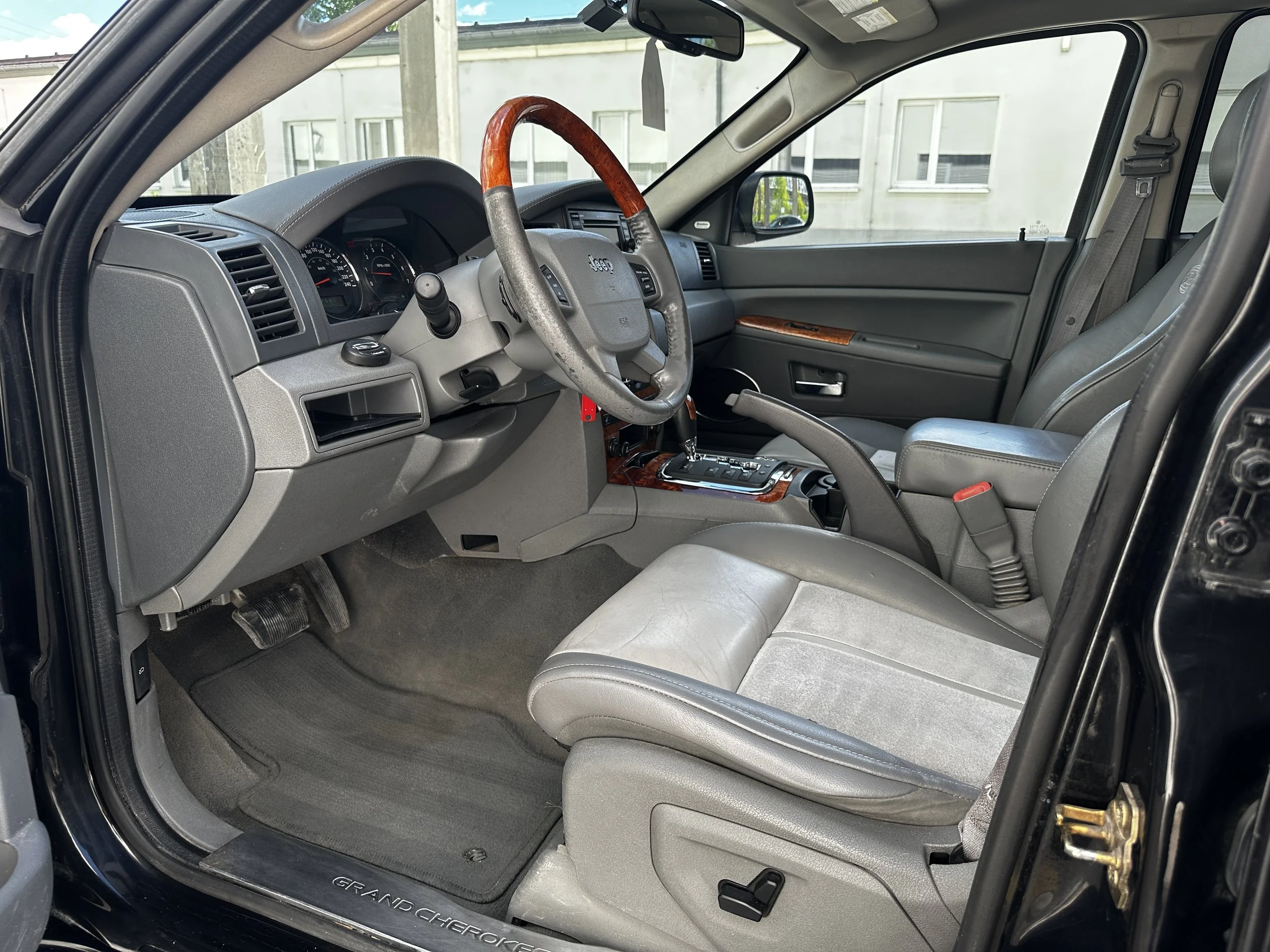 Innenansicht eines Fahrzeugs, Fokus auf das Armaturenbrett, Lenkrad mit Holzgriff, Leder- und Plastikmaterialien, Automatikgetriebe, Fahrersitz und Fußraum sichtbar.