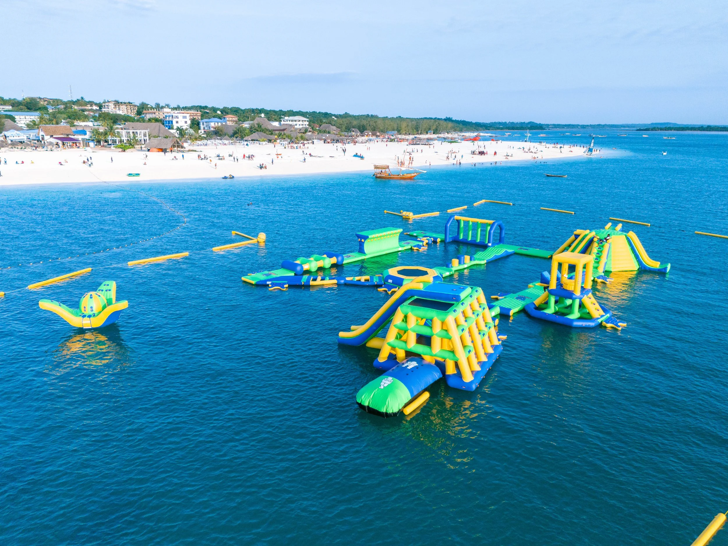 Colorful inflatable water park with slides and climbing structures on a lake, with a sandy beach and houses in the background.
