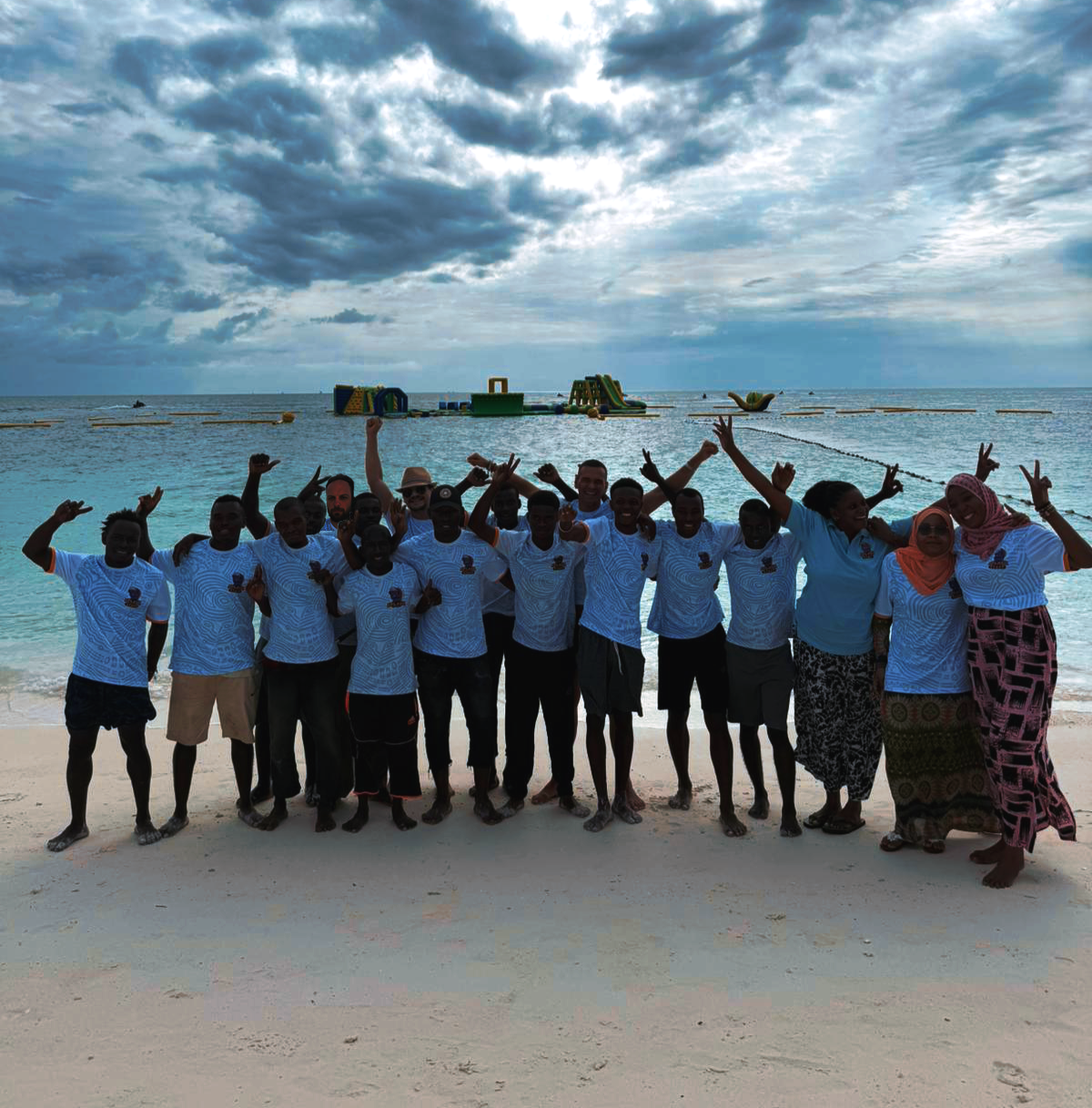 Group of people in matching shirts standing on a beach with inflatable water slides in the background and cloudy sky above.