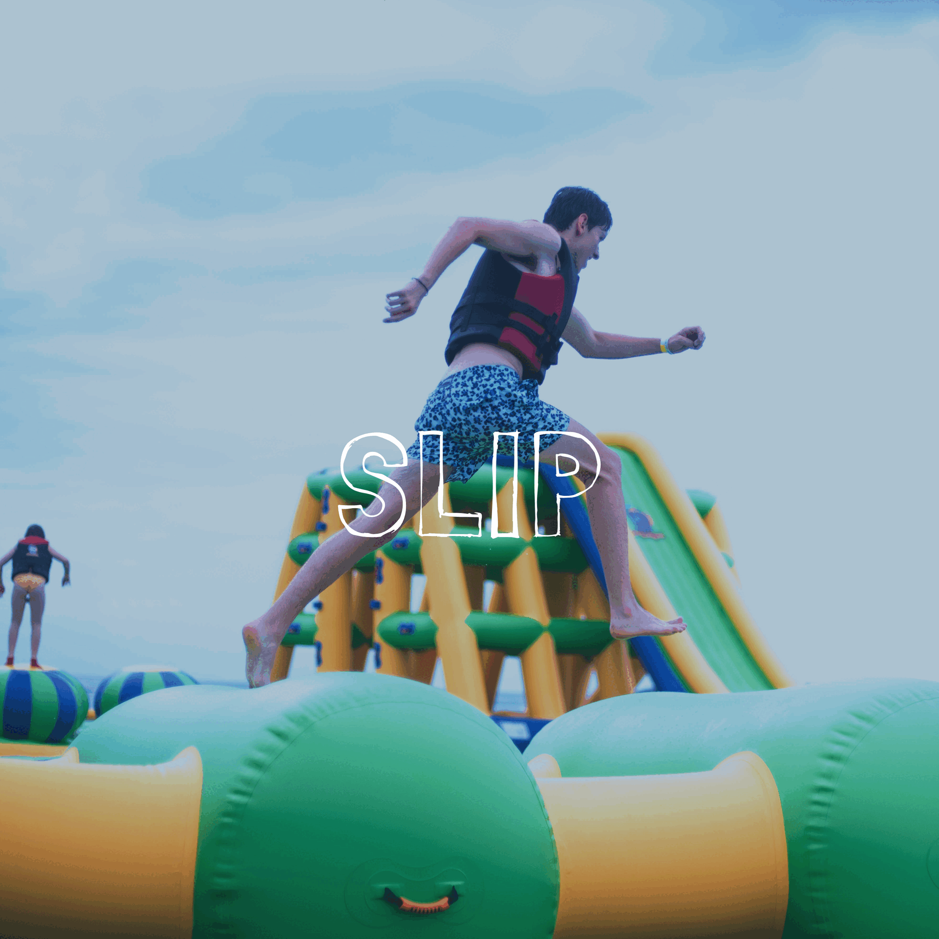 Person jumping on inflatable water obstacle course, wearing a life jacket and swimming shorts, with another person in the background also on the inflatable, cloudy sky overhead.