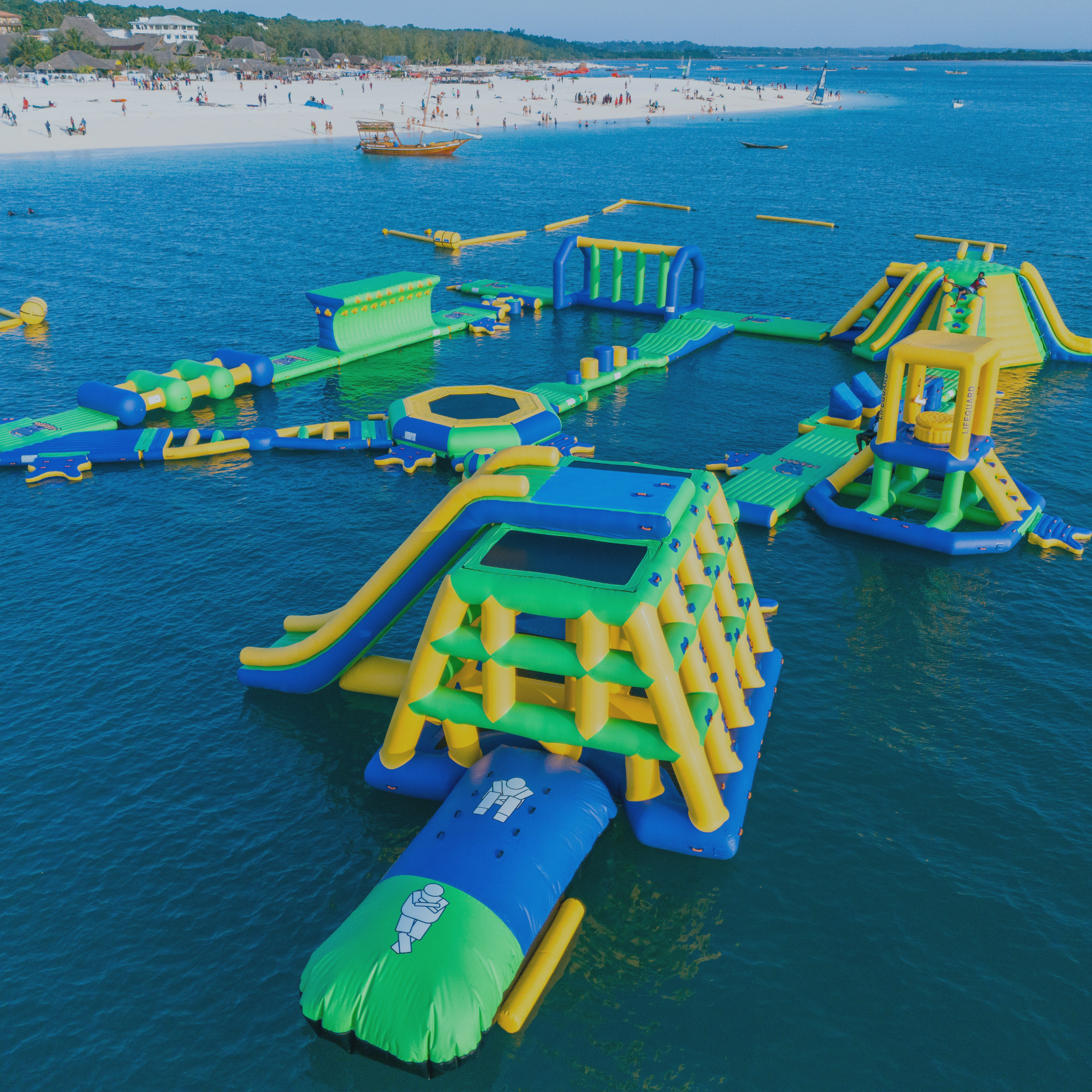 Colorful inflatable water park with slides, climbing structures, and other play areas in a lake, with a crowded white sandy beach and people in the background.
