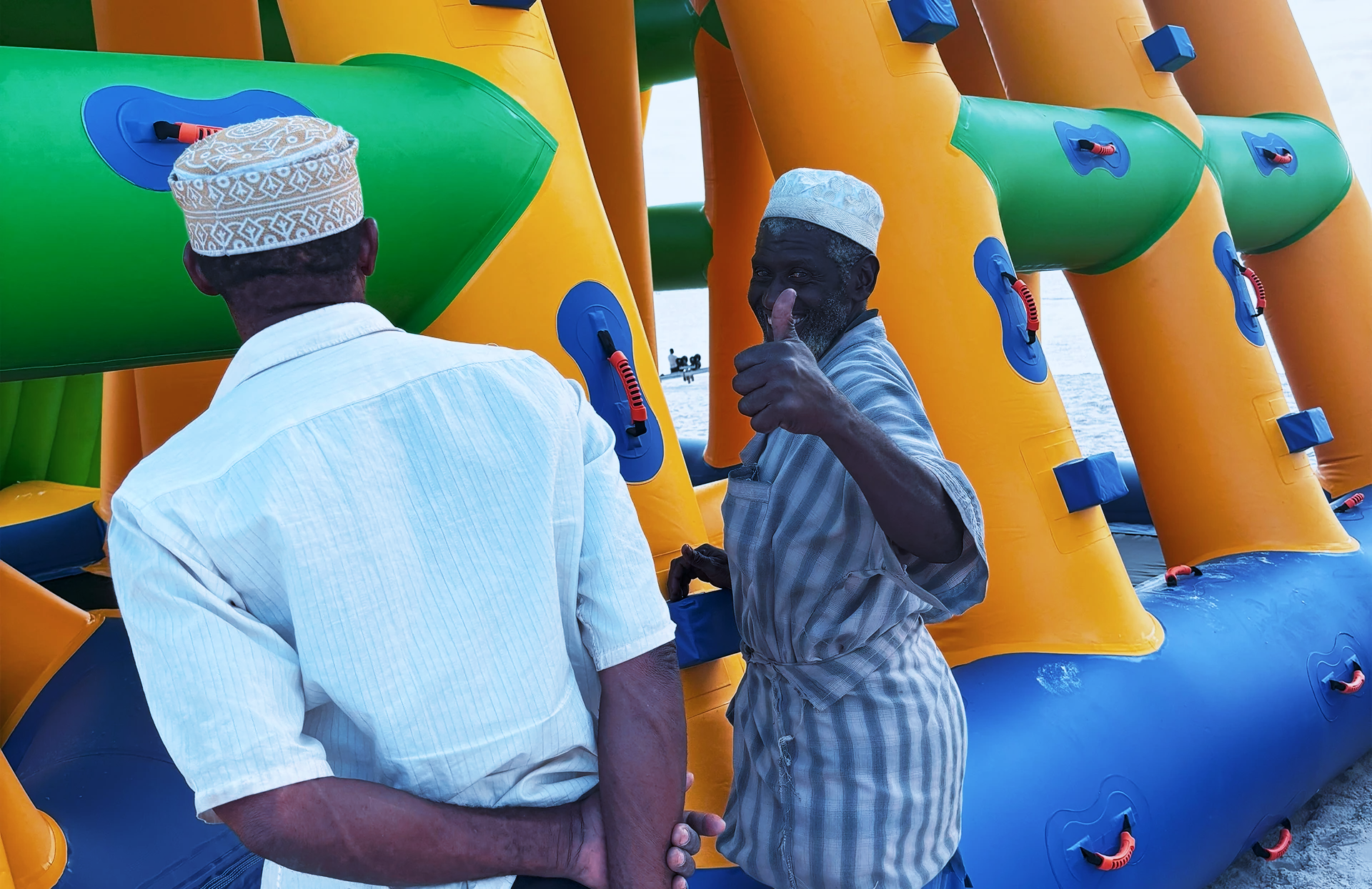 Two elderly men in traditional hats and light clothing standing near a colorful inflatable water slide at the beach. One man is giving a thumbs-up and smiling at the camera.