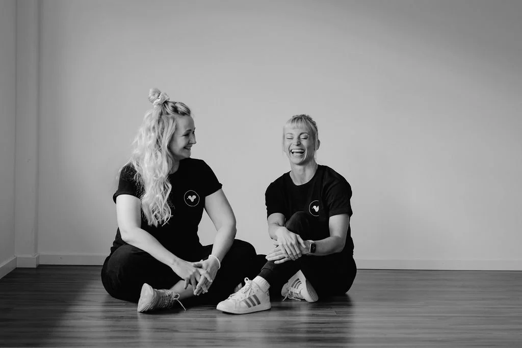Two women sitting on a wooden floor, smiling and laughing while wearing matching black t-shirts, in front of a plain wall.