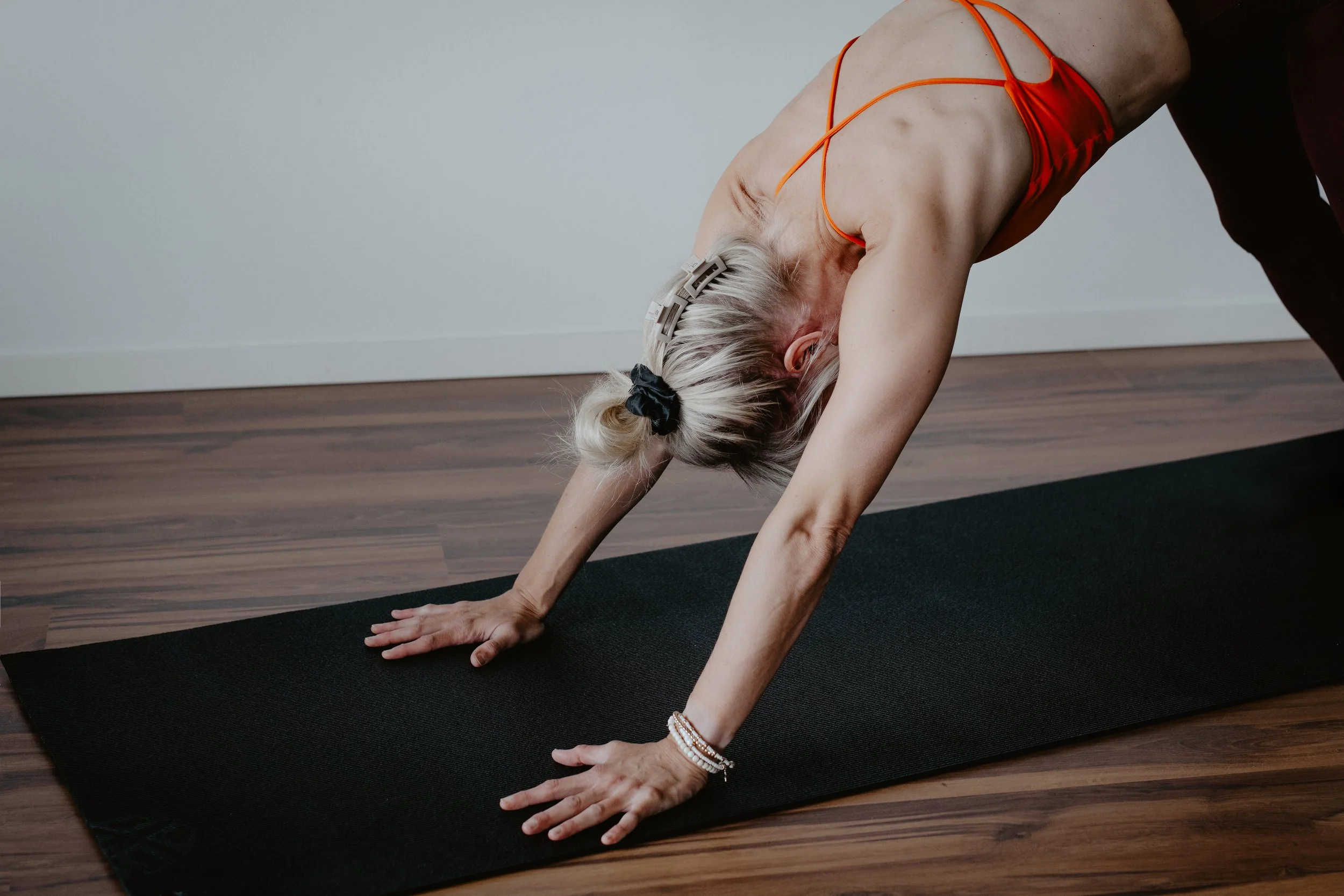 An elderly woman with gray hair in hair clips practicing yoga on a black mat, bending forward with her hands on the ground inside a room with wood flooring and a white wall.