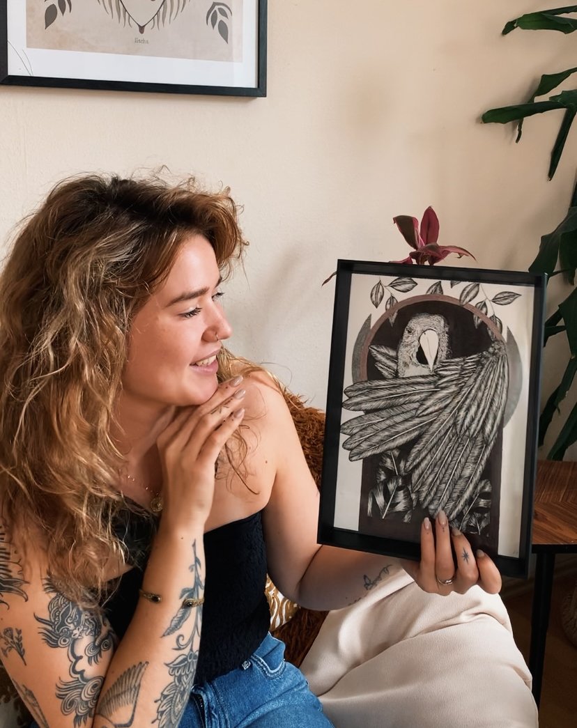 Junge Frau mit Tattoos hält ein gerahmtes Kunstwerk eines Vogelporträts, während sie lächelnd schaut.