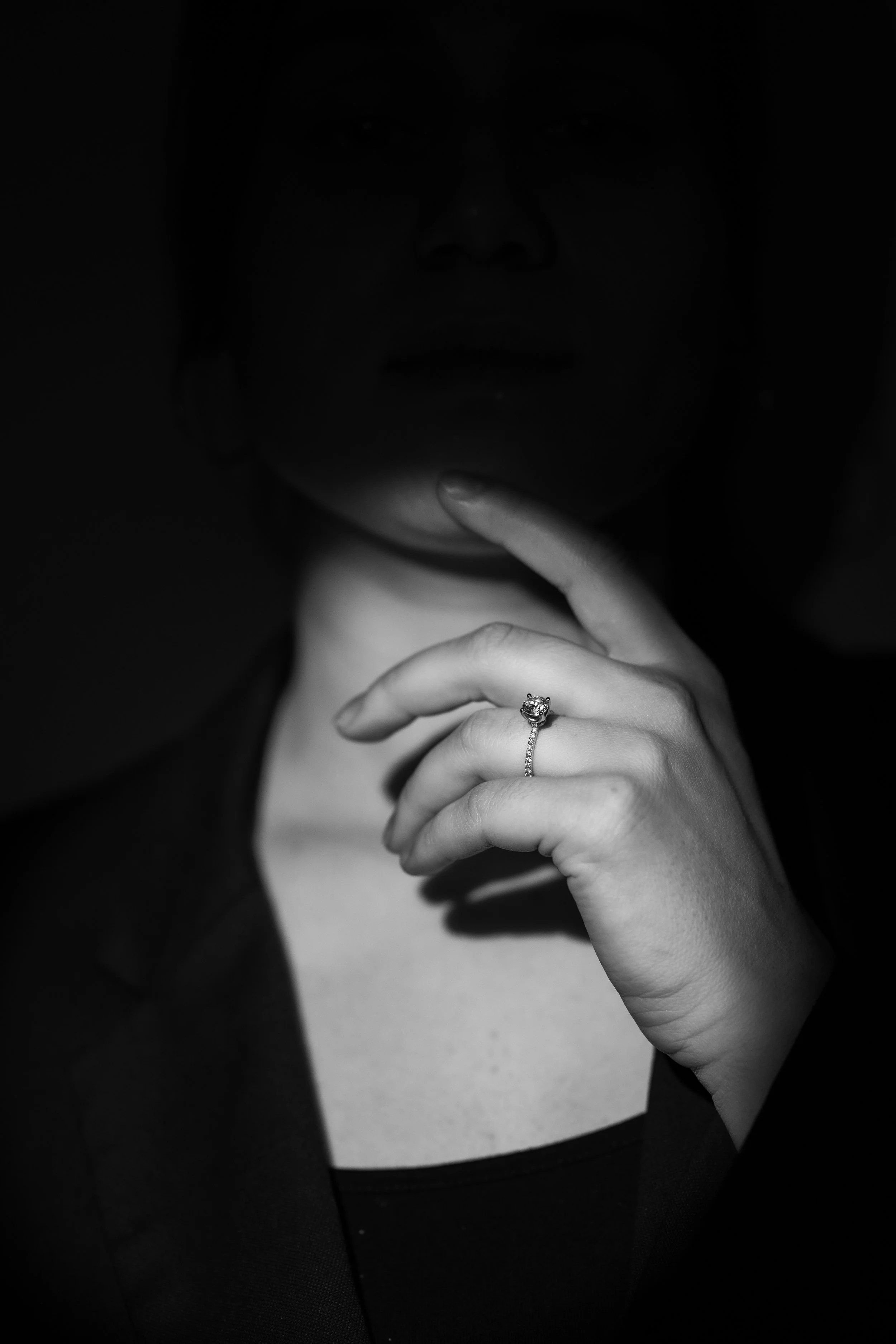 Schwarz-Weiß-Fotografie einer Person, die eine Hand mit einem Ring am Finger auf die Lippen legt, das Gesicht der Person ist kaum sichtbar.