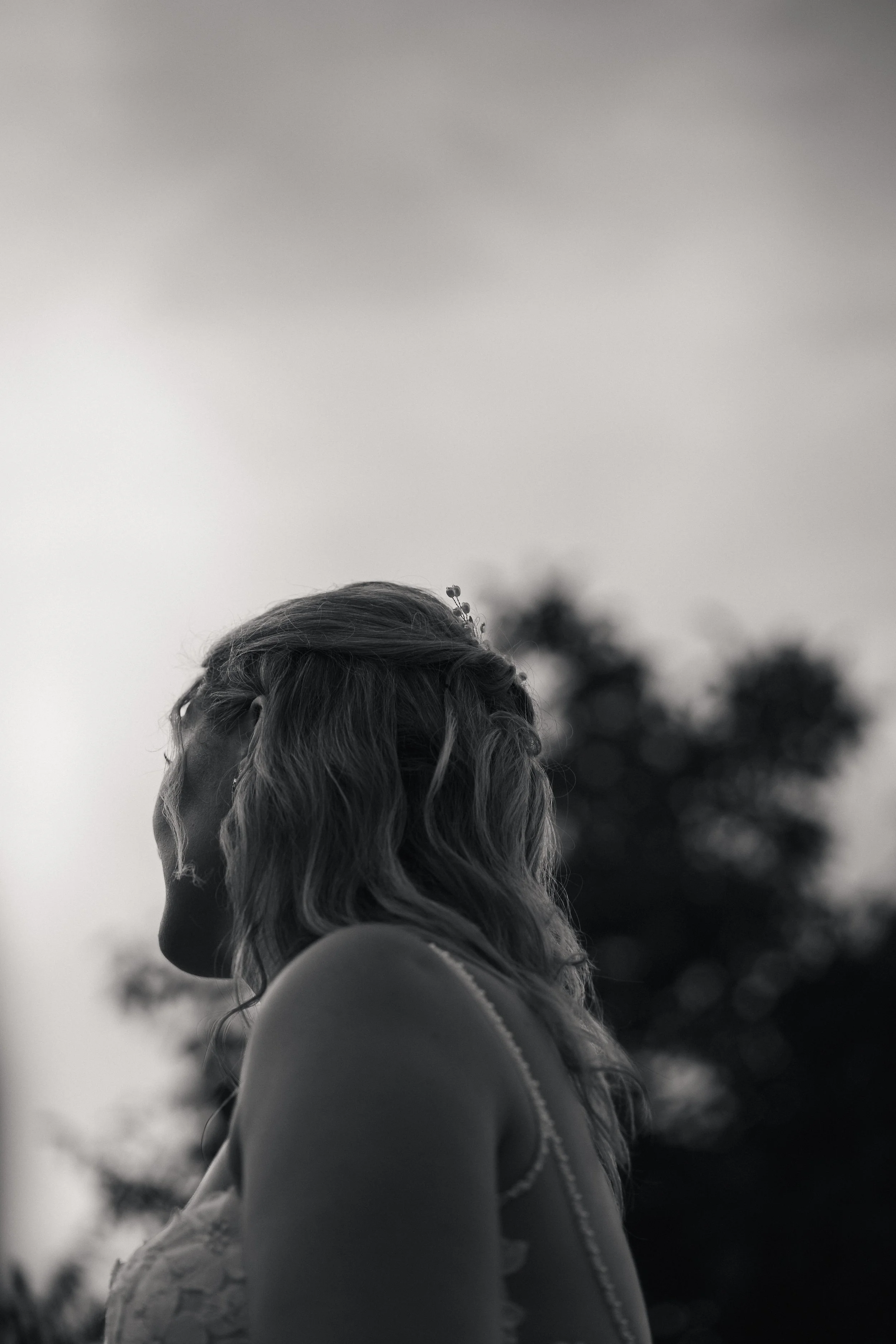 Schwarz-Weiß-Fotografie eines Mädchens mit blonden, gewellten Haaren, das seitlich blickt, im Freien mit Bäumen im Hintergrund.