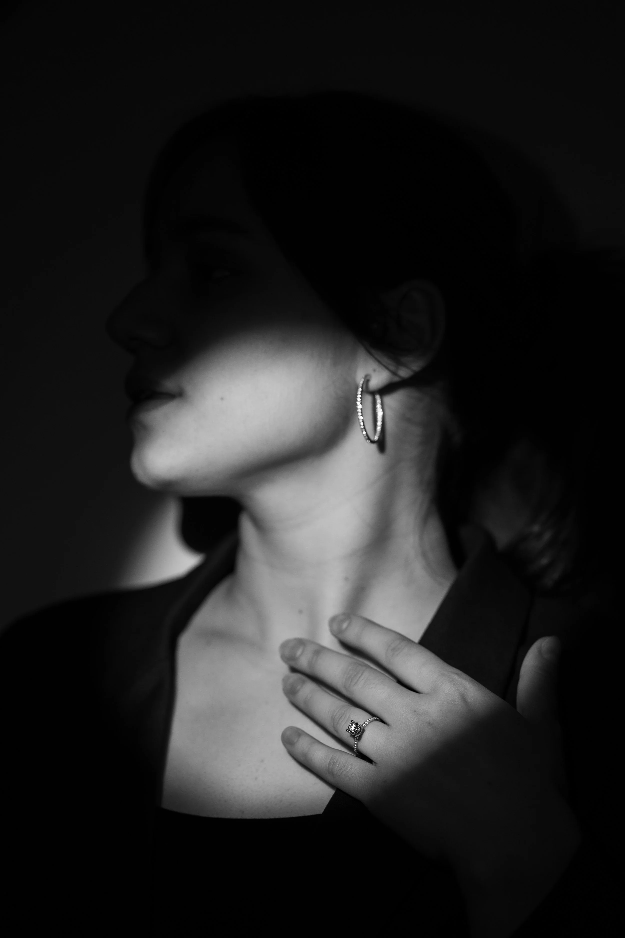 Schwarz-weiße Aufnahme einer Frau in Seitenansicht, die an ihrer Schulter berührt wird, mit Ohrringen und Ehering sichtbar.