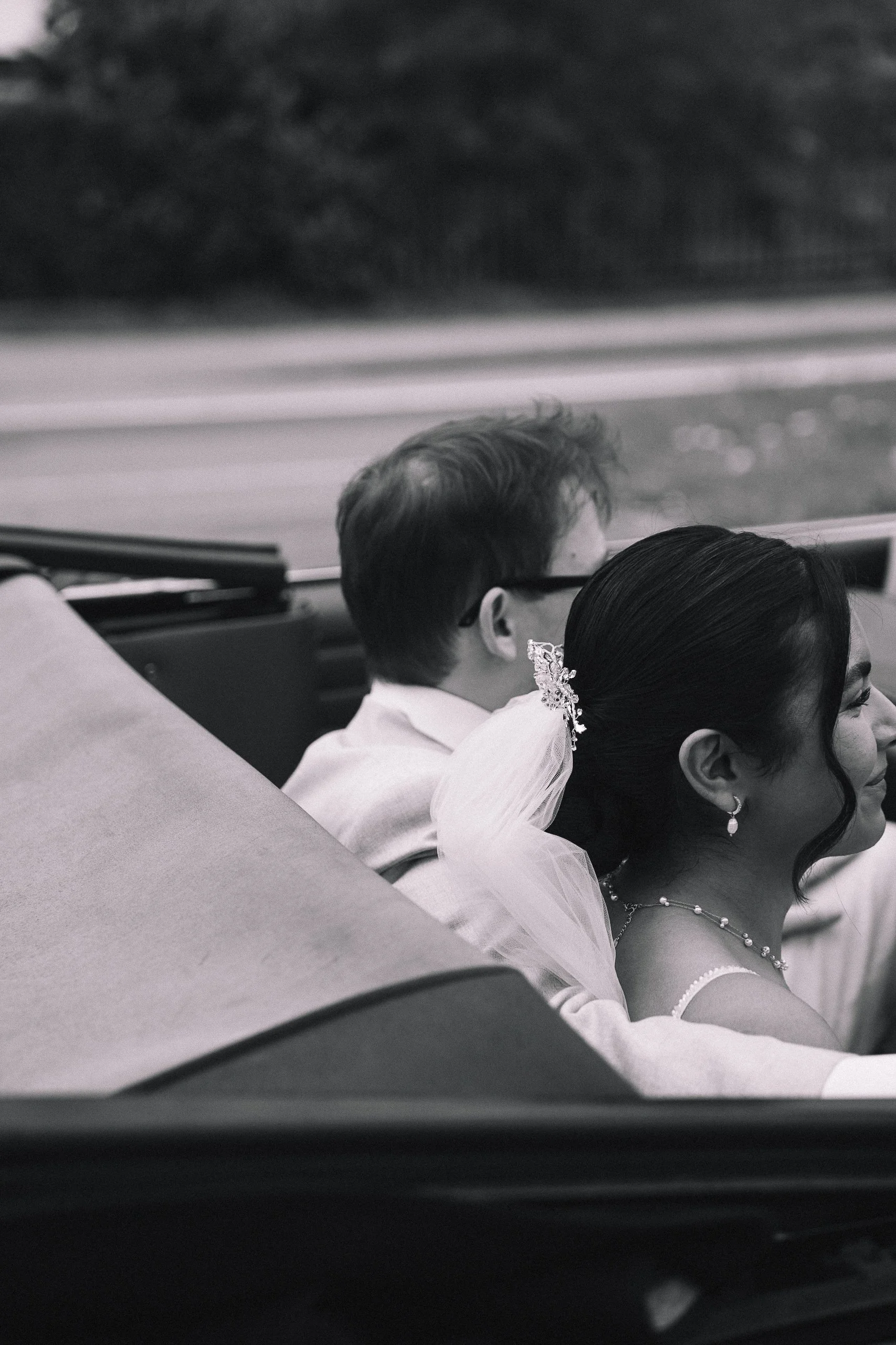 Schwarz-weiß-Foto eines Paares, das eng zusammen in einer Limousine sitzt. Die Frau trägt elegante Ohrringe, ein Haar-Accessoire und ein Abendkleid, während der Mann eine Brille trägt. Sie scheinen sich aneinander zu lehnen, möglicherweise bei einer besonderen Gelegenheit.