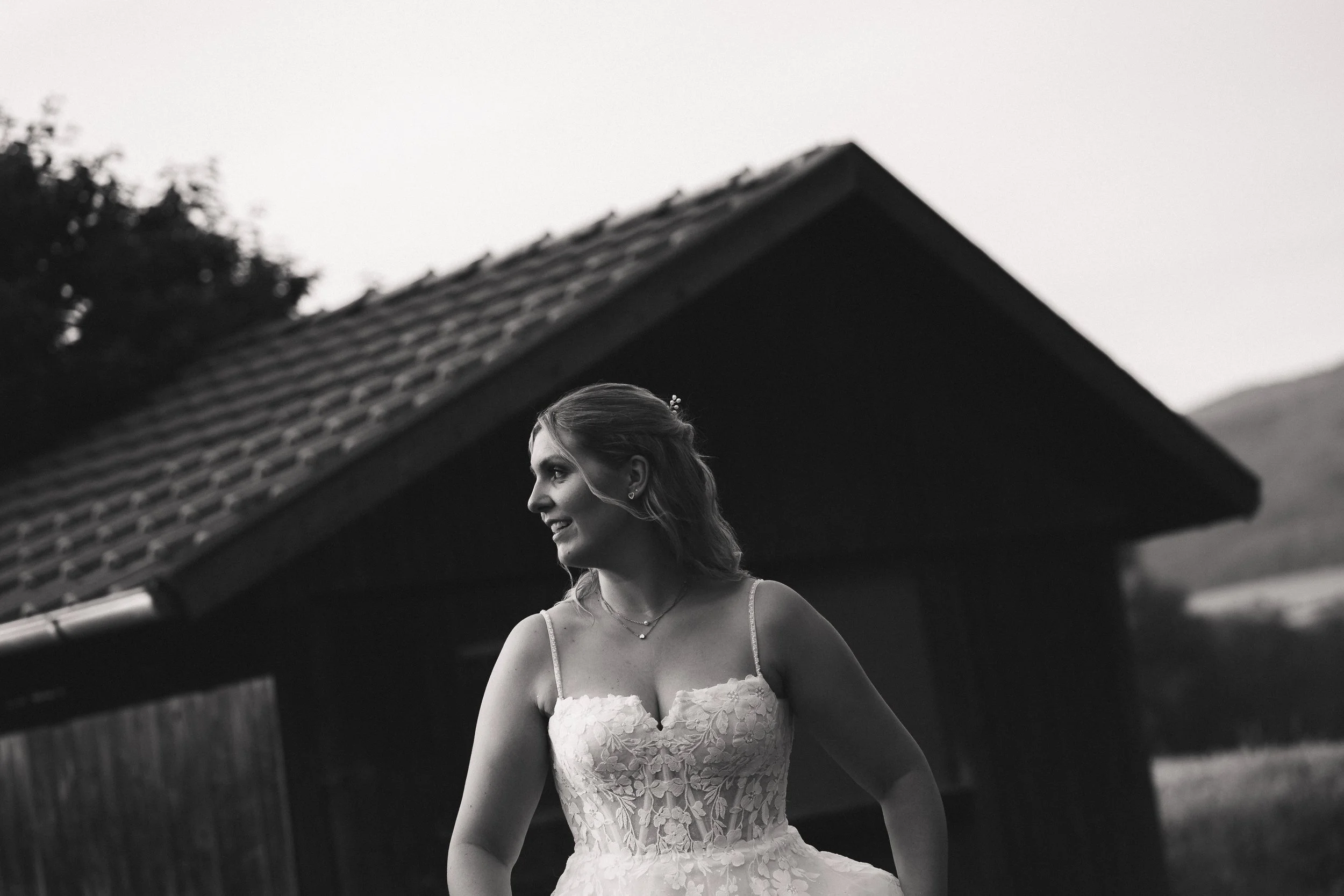 Schwarz-Weiß-Fotografie einer jungen Frau in einem Hochzeitskleid, die lächelt, im Freien neben einem Gebäude mit geneigtem Dach, bei Sonnenuntergang oder Dämmerung.